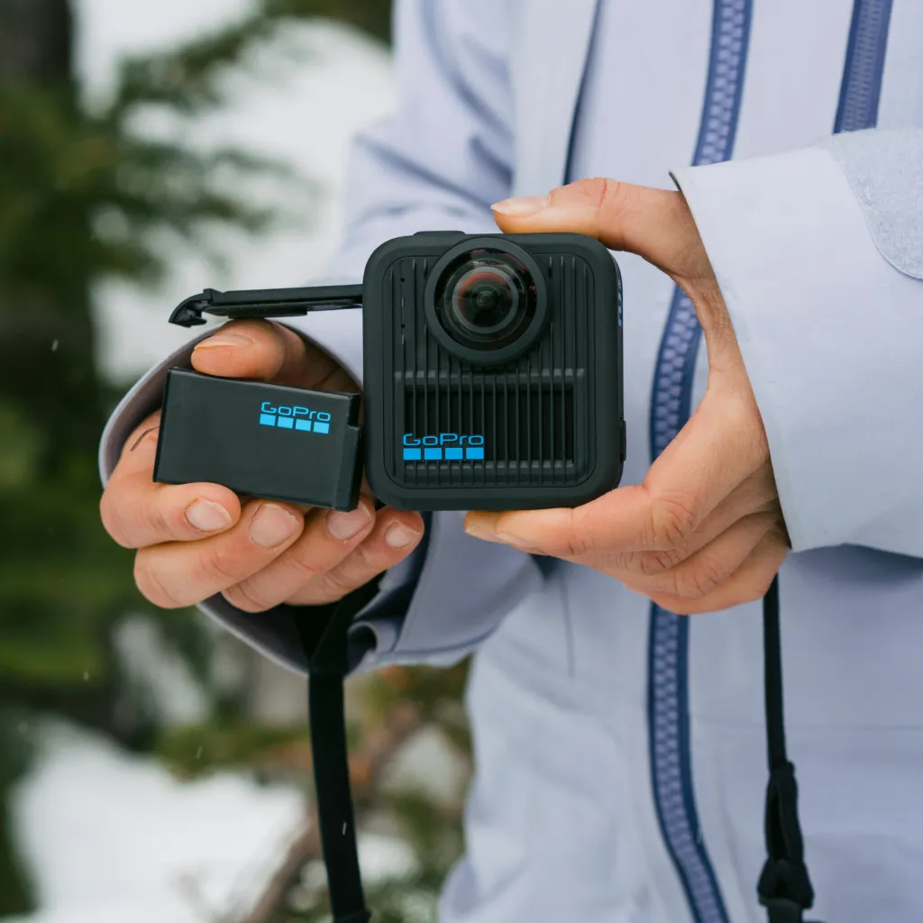 Egy személy egy fekete GoPro kamerát és akkumulátort tart. A kamerán lencse és a GoPro logó látható. Havas háttér.