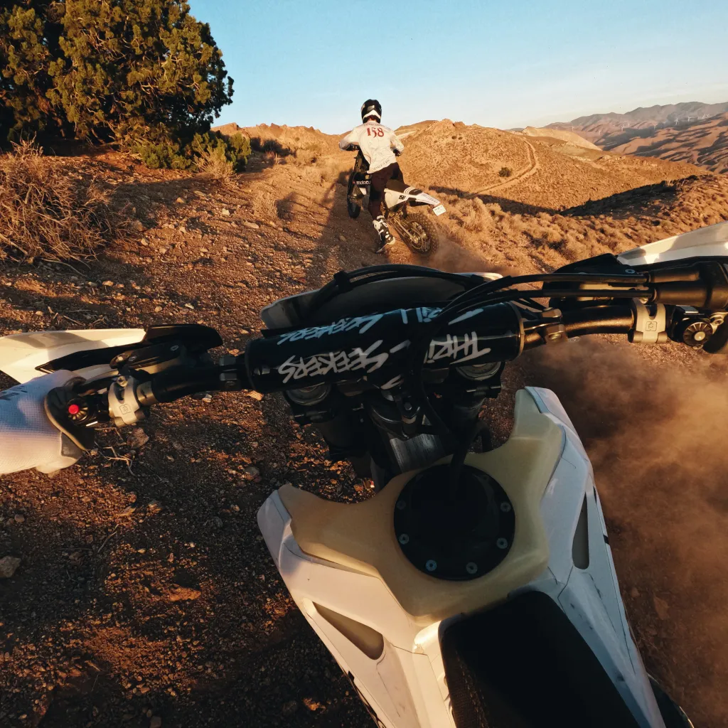 Motoros egy dirt bike-on, a motoros szemszögéből, egy másik motorost követve felfelé egy földúton egy sivatagi tájon.