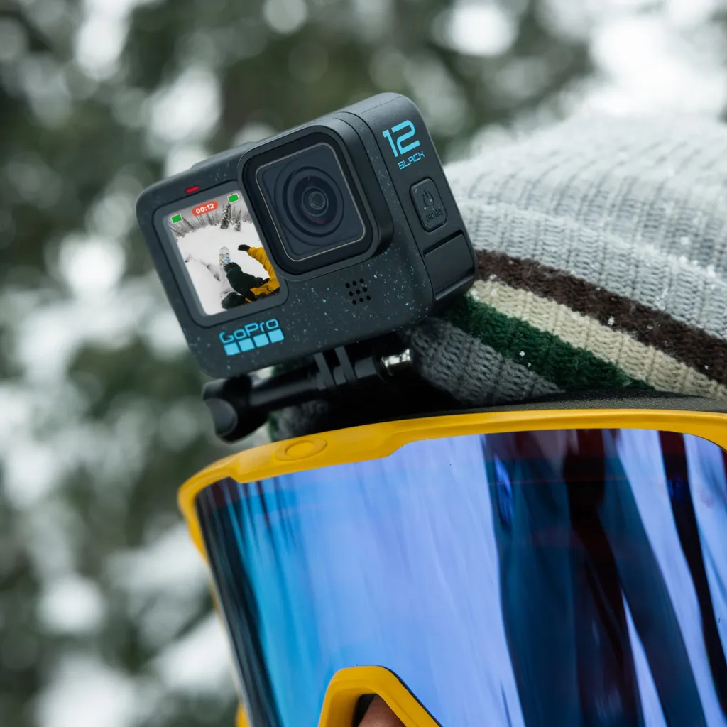 Egy GoPro 12 Black kamera egy személy fejére van szerelve. A személy sí szemüveget és szürke sapkát visel. Hó van a háttérben.