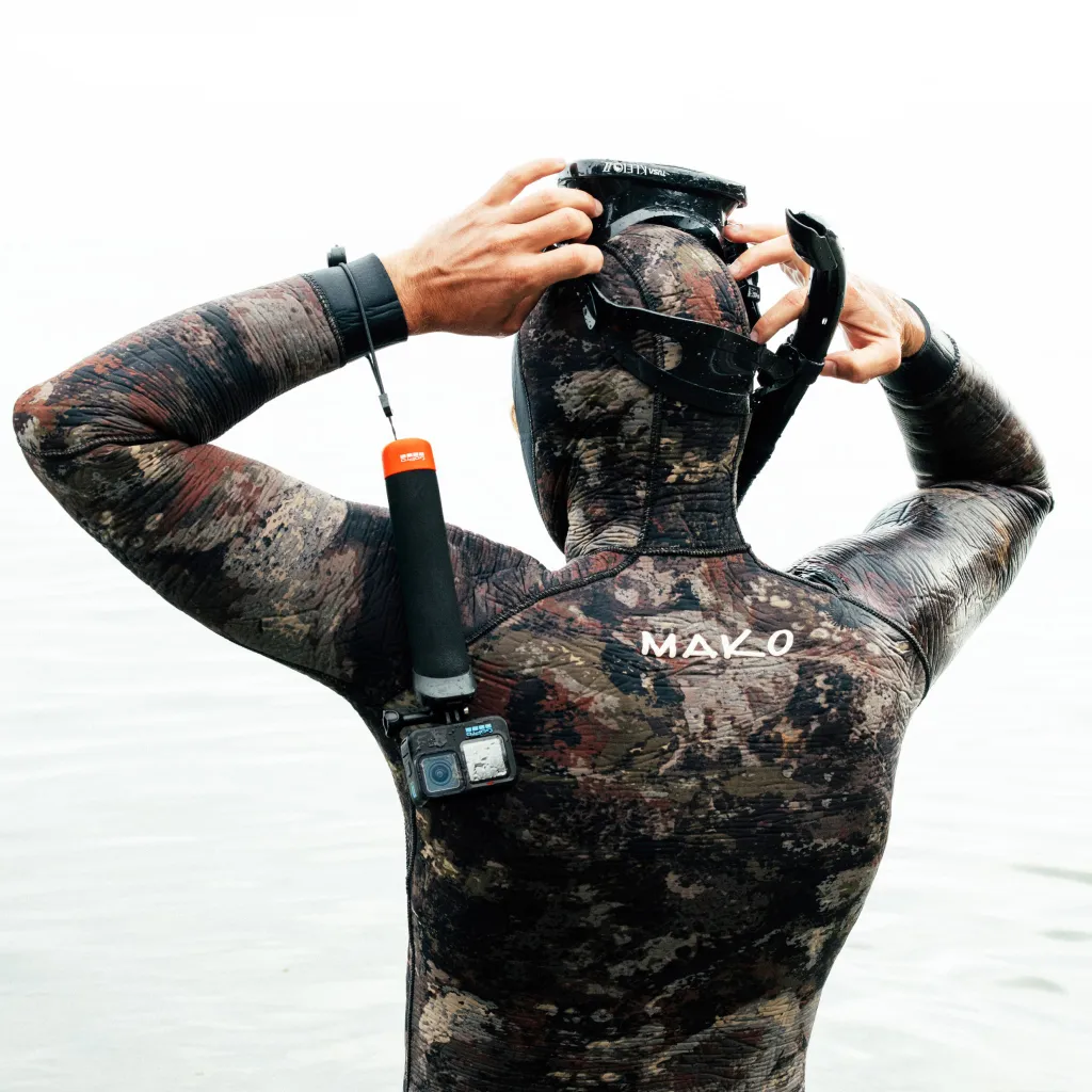 A búvárruhás személy a búvármaszkot és a snorkelt igazítja. Kamera csatolva. A búvárruha álcázó mintázatú. A 'MAKO' szó látható.