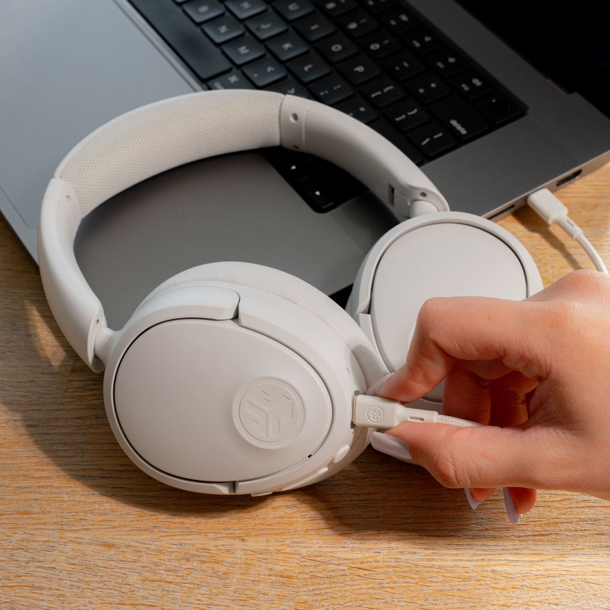 Weisse Kopfhörer auf einem Laptop. Eine Hand steckt das Ladekabel ein. Holztisch im Vordergrund.