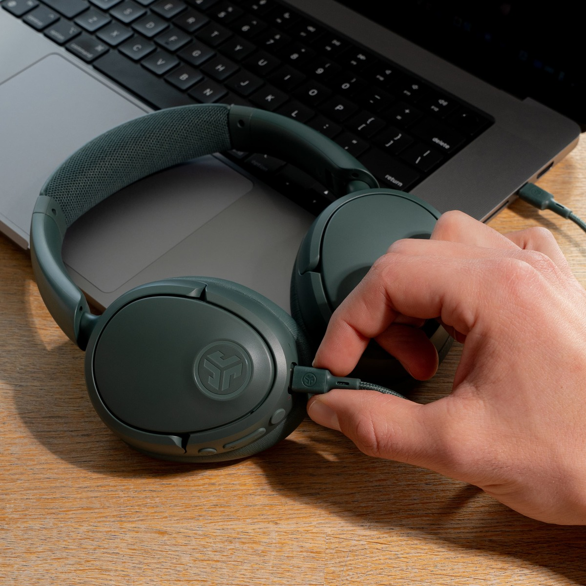 Grüne Kopfhörer liegen auf einem Laptop. Eine Hand steckt ein Kabel ein. Holzoberfläche.