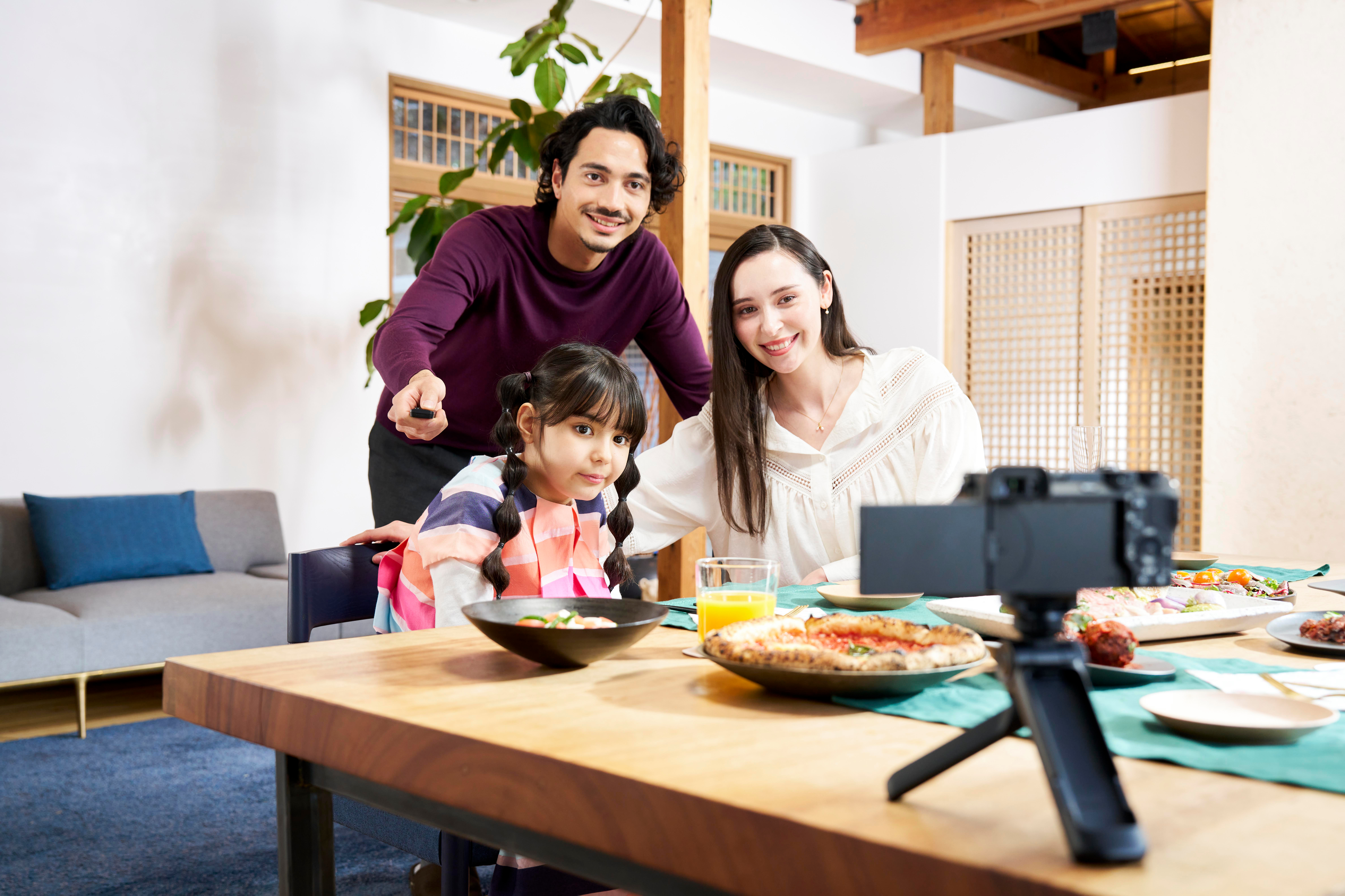 Eine dreiköpfige Familie lächelt in die Kamera. Ein Tisch ist mit Essen und einer Kamera gedeckt. Ein Mann hält eine Fernbedienung.