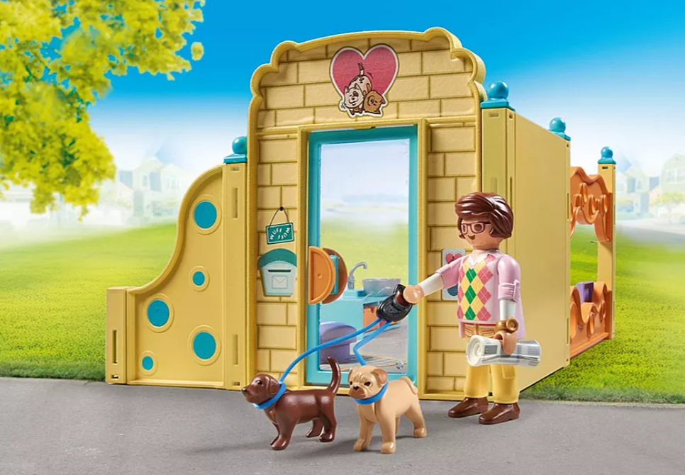 Eine Playmobil-Tierklinik, in der eine Person mit zwei Hunden spazieren geht. Die Klinik ist gelb mit einem Herzschild. Grünes Gras und blauer Himmel sind im Hintergrund zu sehen.