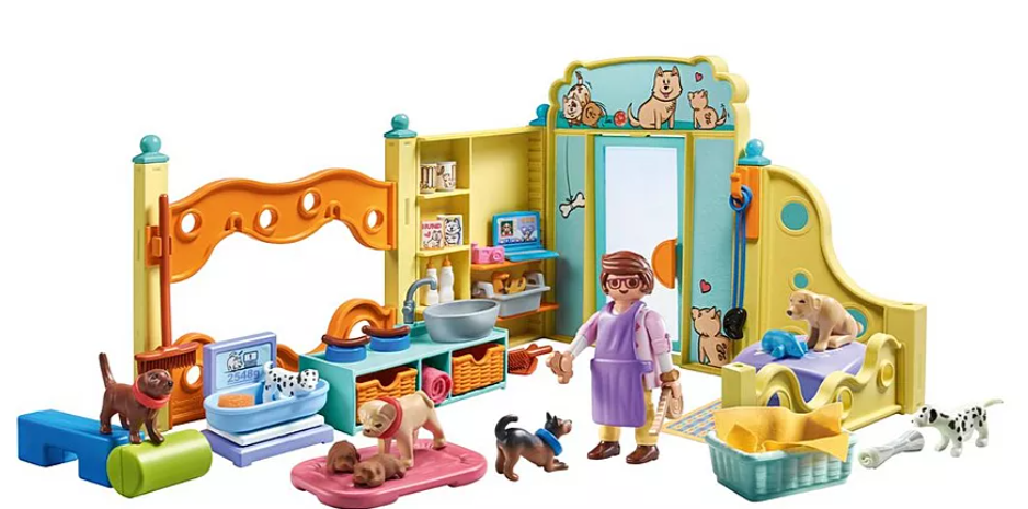 Ein Spielset für Hundefriseur. Es enthält eine Frauenfigur, mehrere Spielzeughunde, eine Pflegestation, Betten und Zubehör. Die Szene ist in Innenräumen.