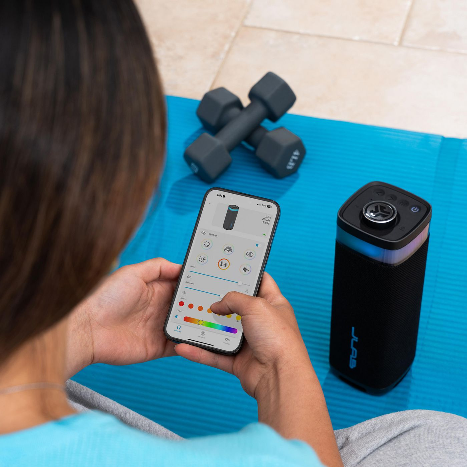Een persoon gebruikt een smartphone om een luidspreker te bedienen. Halters en een blauwe yogamat op de achtergrond.