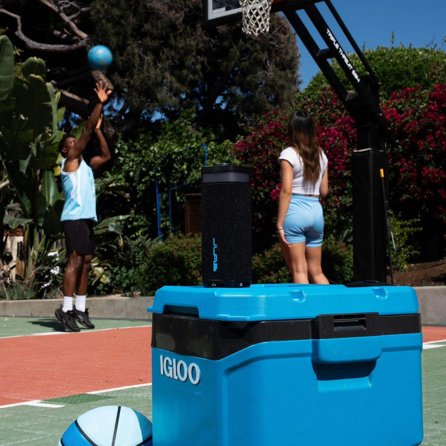 Een man schiet een blauwe basketbal. Een vrouw kijkt toe. Een koeler, speaker en bal staan op de voorgrond. Een basketbalring staat op de achtergrond.