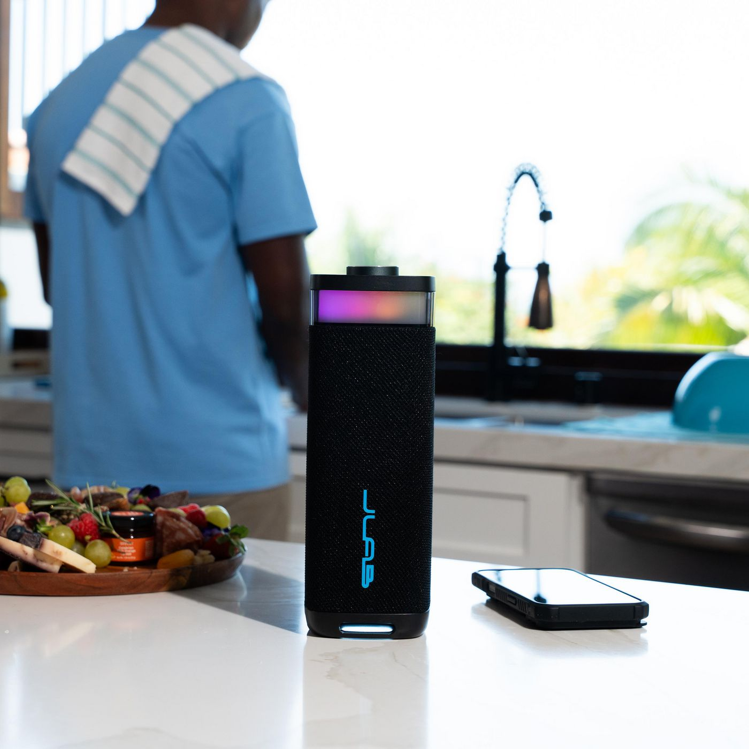 Een zwarte luidspreker met blauw logo staat op een wit aanrecht. Een persoon is op de achtergrond. Een telefoon en eten zijn ook aanwezig.