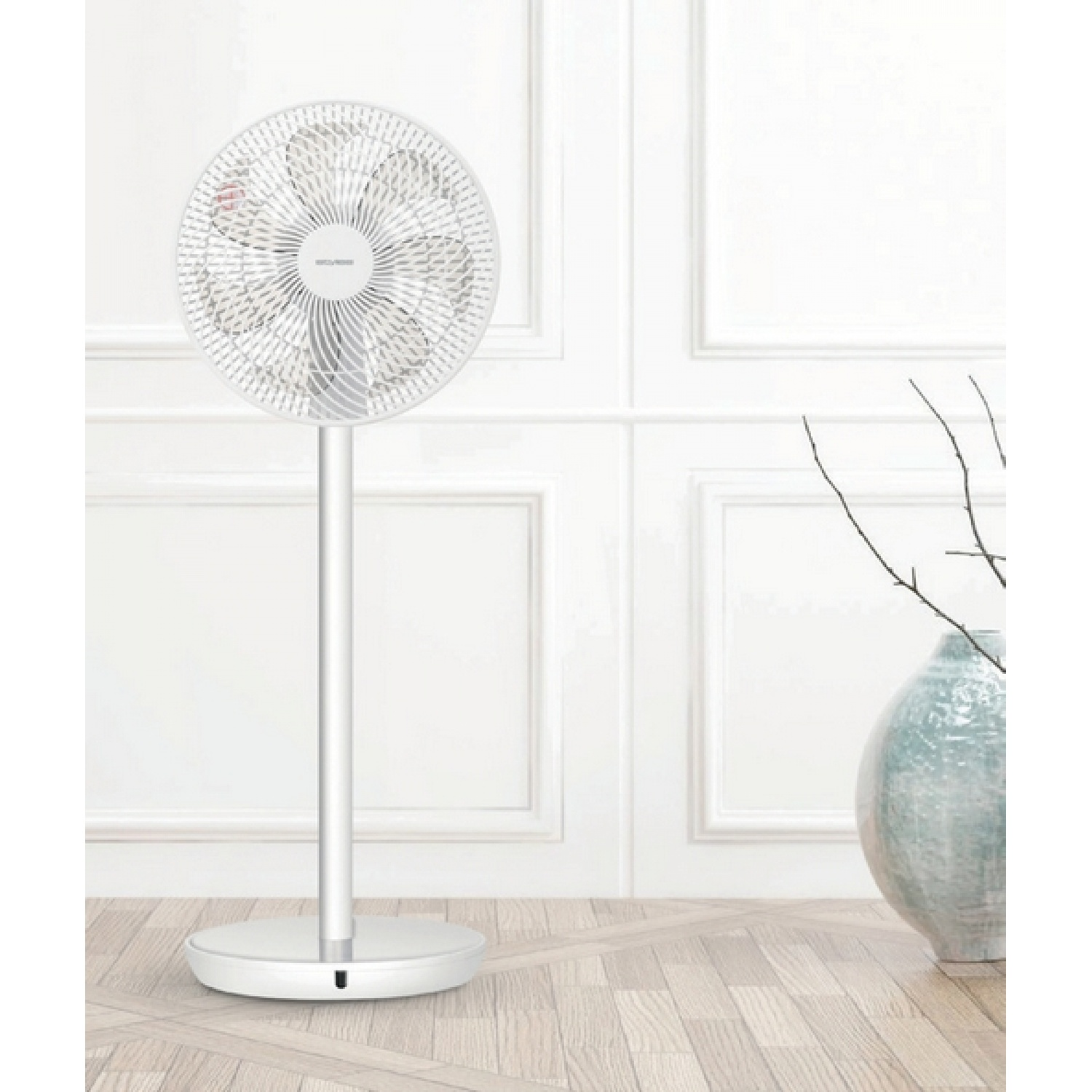 Un ventilateur sur pied blanc est dans une pièce. Un vase avec des branches est sur un sol en bois, contre un mur blanc.