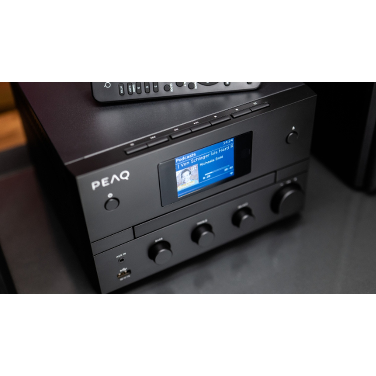 Un sistema audio PEAQ nero con un telecomando sopra. Lo schermo mostra un podcast e l'ora. Sono visibili manopole e pulsanti.