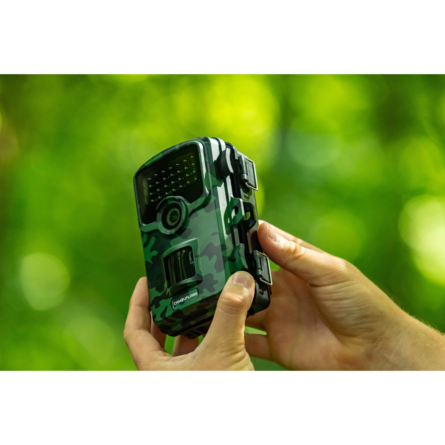 Una persona tiene in mano una fotocamera da caccia mimetica con entrambe le mani. Lo sfondo mostra una foresta verde sfocata.