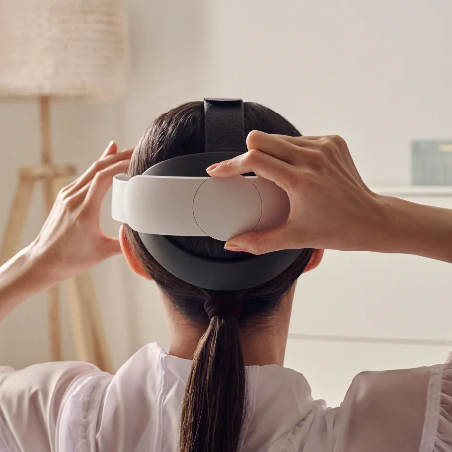 Una persona sta indossando un visore VR. L'auricolare è bianco e nero. Indossa una camicia bianca. Lo sfondo è una stanza luminosa.