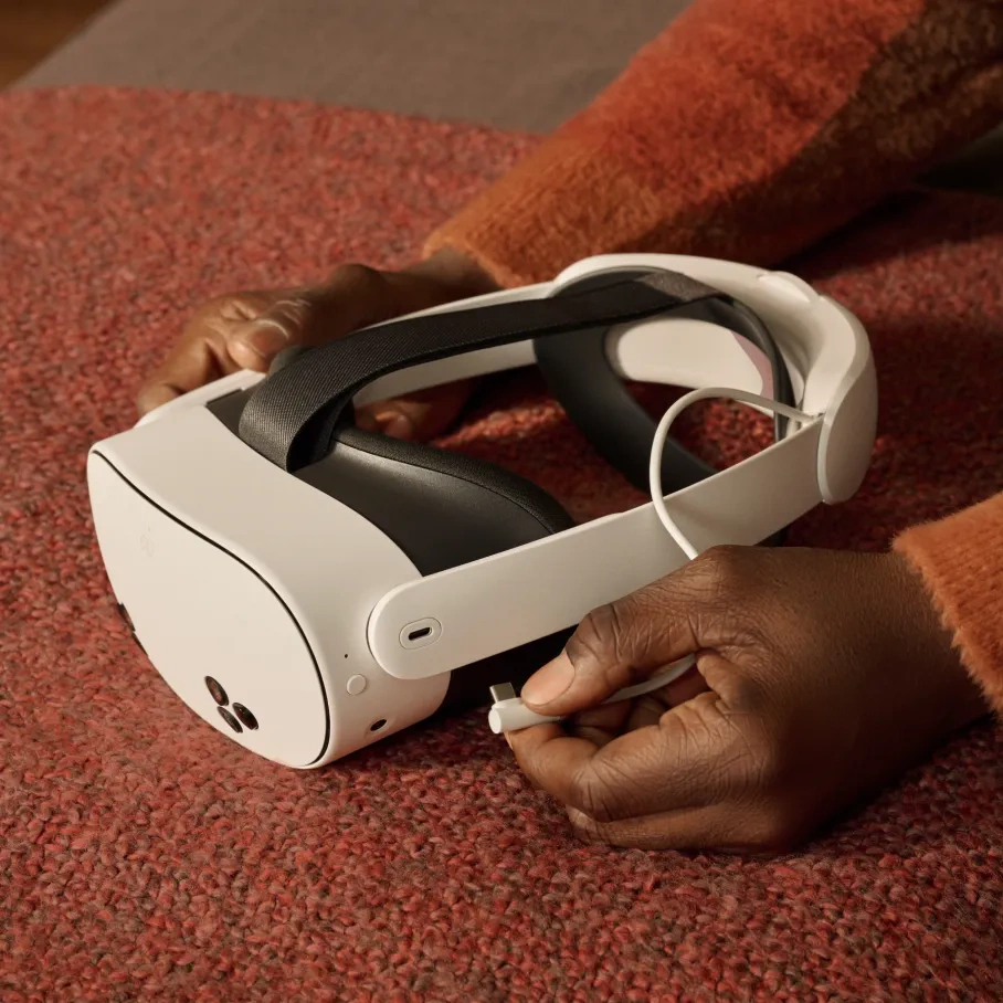 Hände halten ein weißes VR-Headset mit schwarzem Riemen und weißem Kabel. Die Person trägt einen orangefarbenen Pullover.