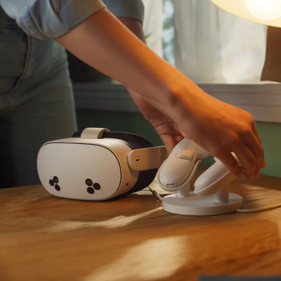 Una mano posiziona un controller VR bianco su un supporto bianco. Un visore VR bianco è appoggiato su un tavolo di legno.