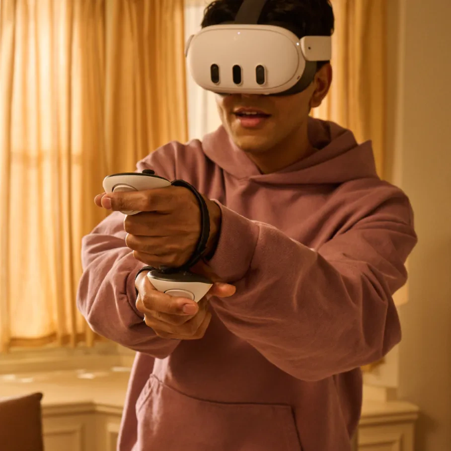 Mann mit VR-Headset und Controllern. Er ist in einem Raum mit Vorhängen und einem Fenster. Er trägt einen rosa Hoodie.