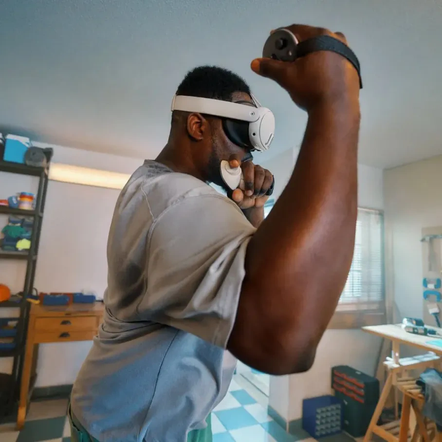 Ein Mann mit VR-Brille trainiert mit Controllern. Er befindet sich in einem Raum mit Regal, Tisch und Fenster. Er boxt.