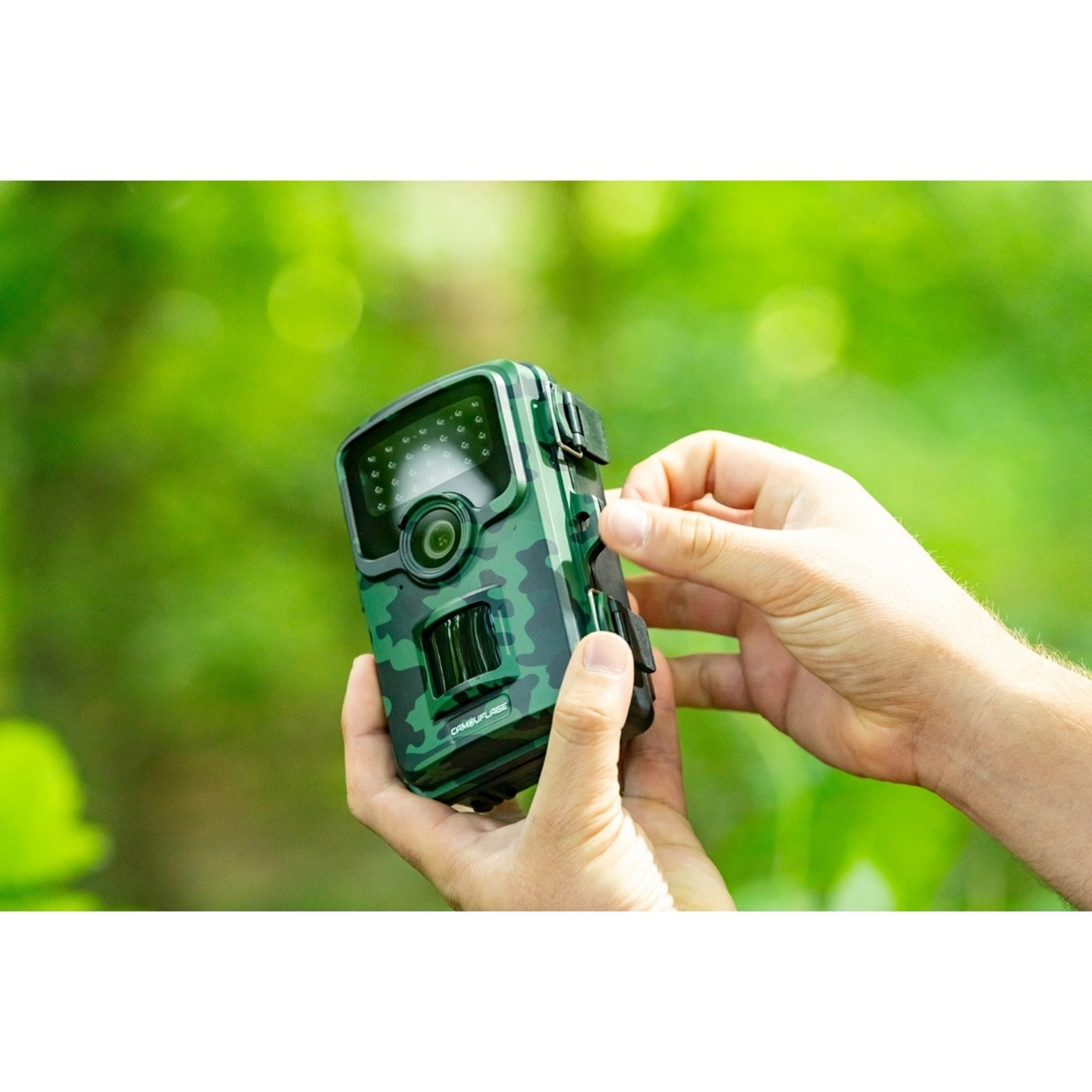 Una persona tiene in mano una fotocamera mimetica. Lo sfondo è un verde sfocato, che indica un ambiente forestale. La fotocamera viene maneggiata.