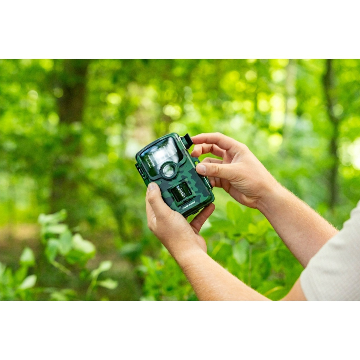 Una persona tiene in mano una fotocamera mimetica in una foresta. La fotocamera è verde. Lo sfondo è un fogliame verde sfocato.