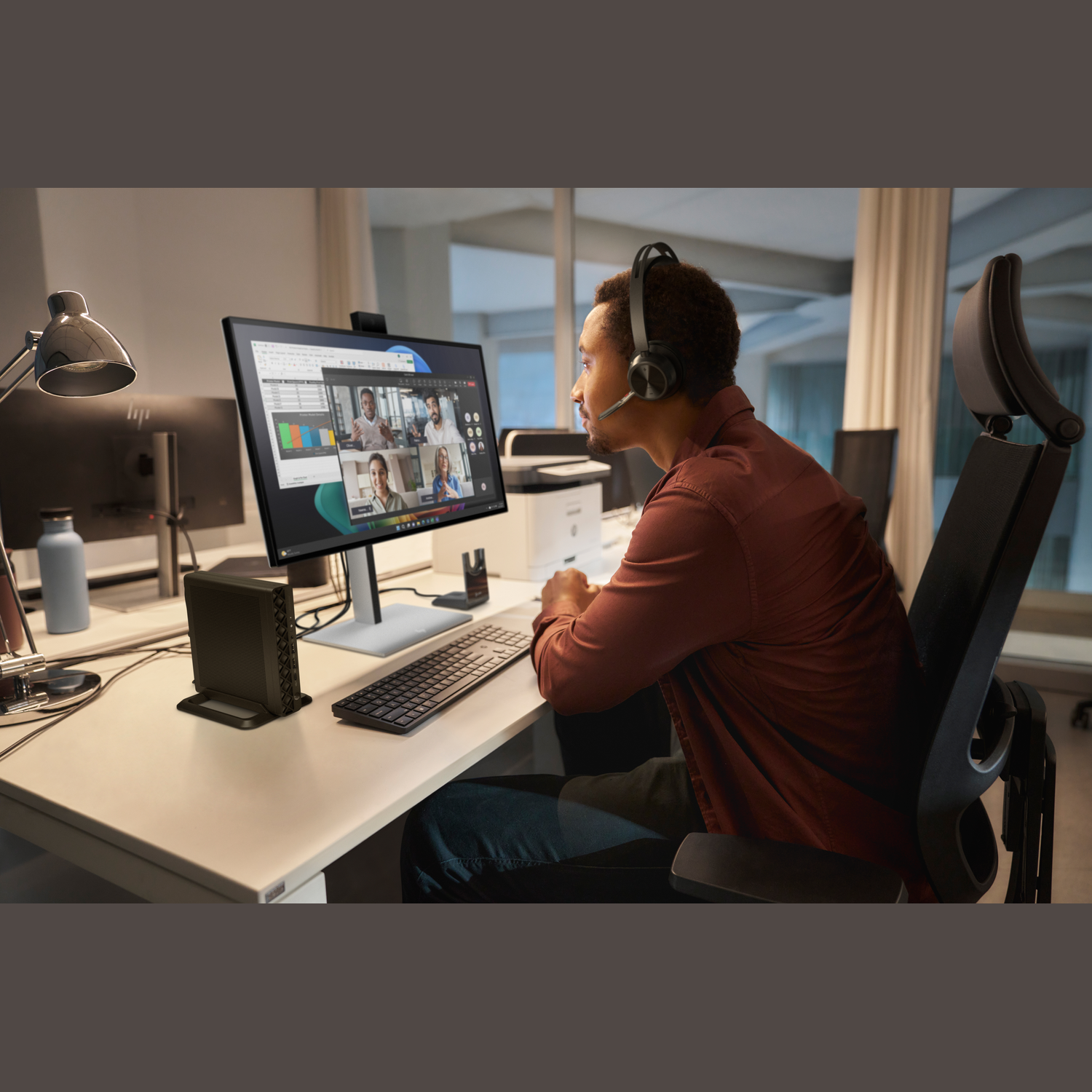 A man wearing a headset sits at a desk with a monitor displaying a video call. Desk has keyboard, speakers and a lamp.