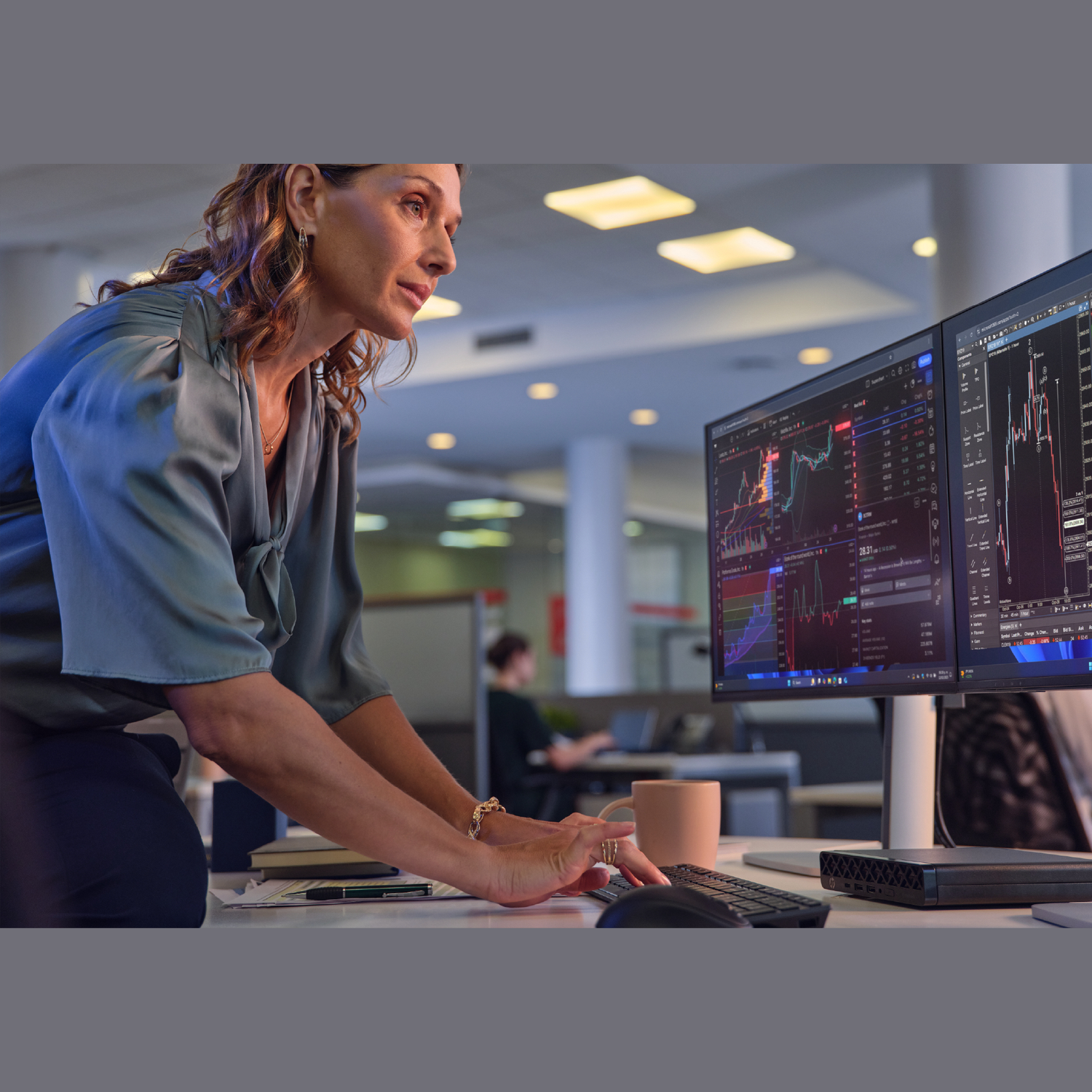 A woman works at a desk with dual monitors displaying stock charts. She wears a blue-green top, with a cup and keyboard on the desk.