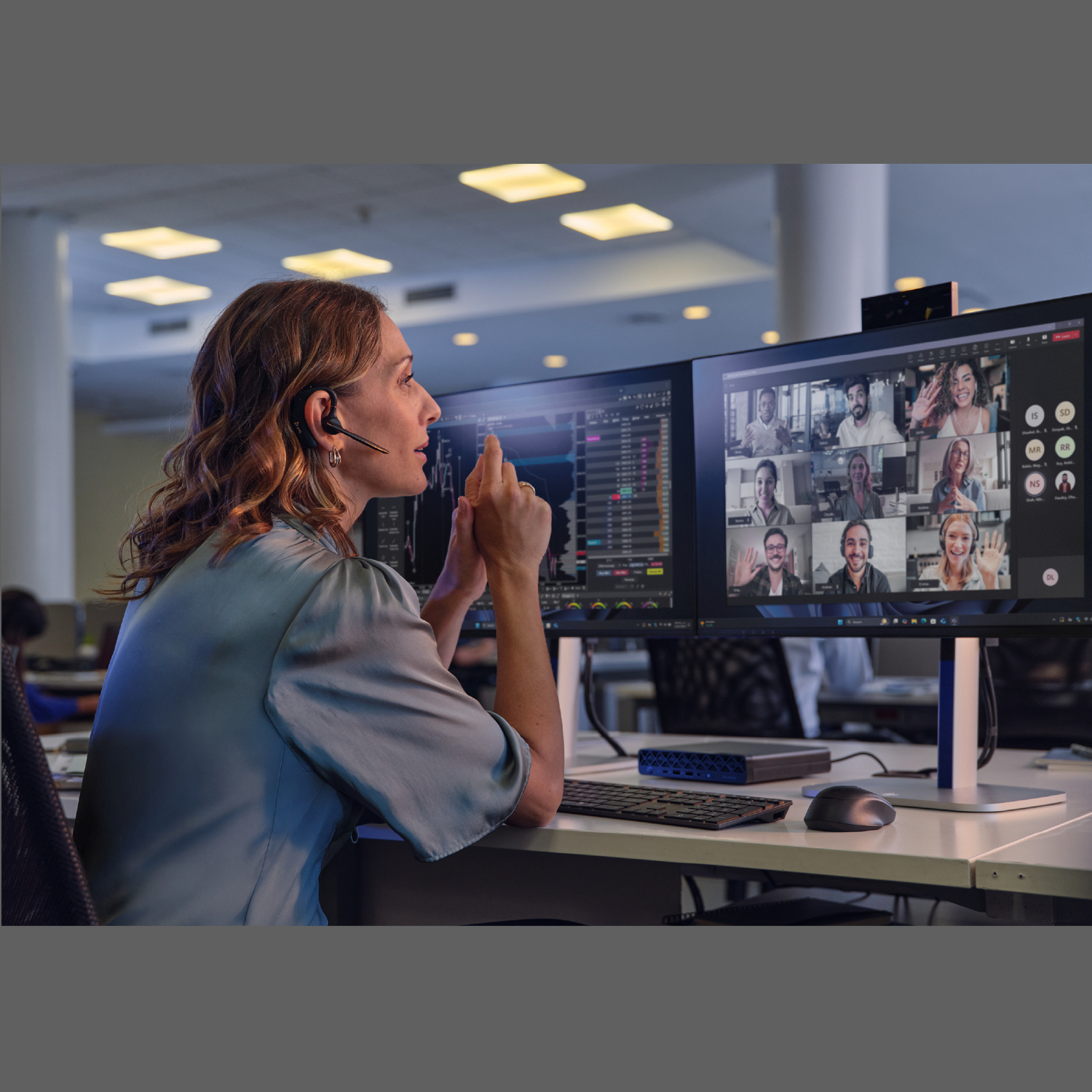 A woman in a headset gestures at a monitor displaying a video call with multiple participants.