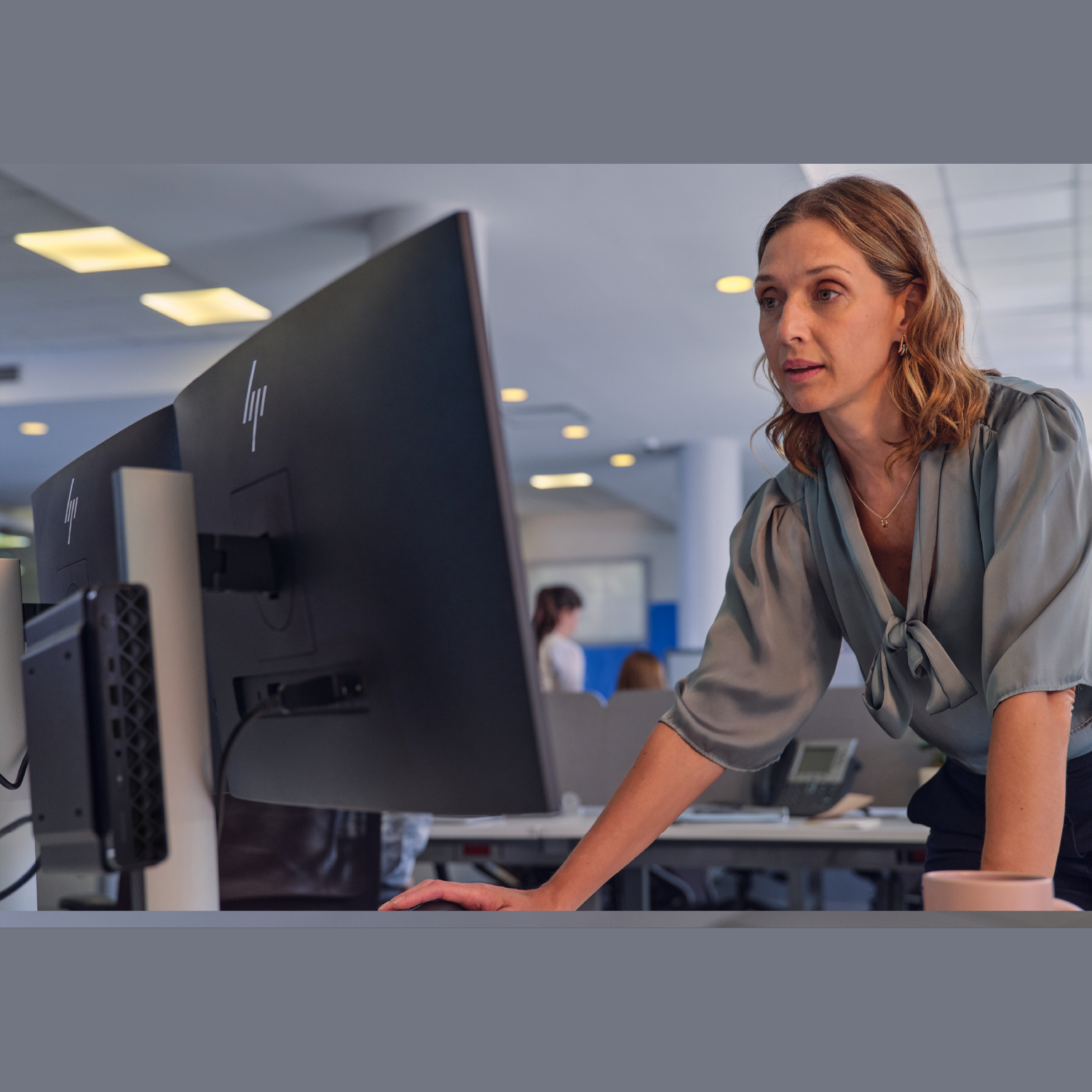 Woman at desk looking at computer screen. She wears a blue blouse. Office environment, other people in the background.