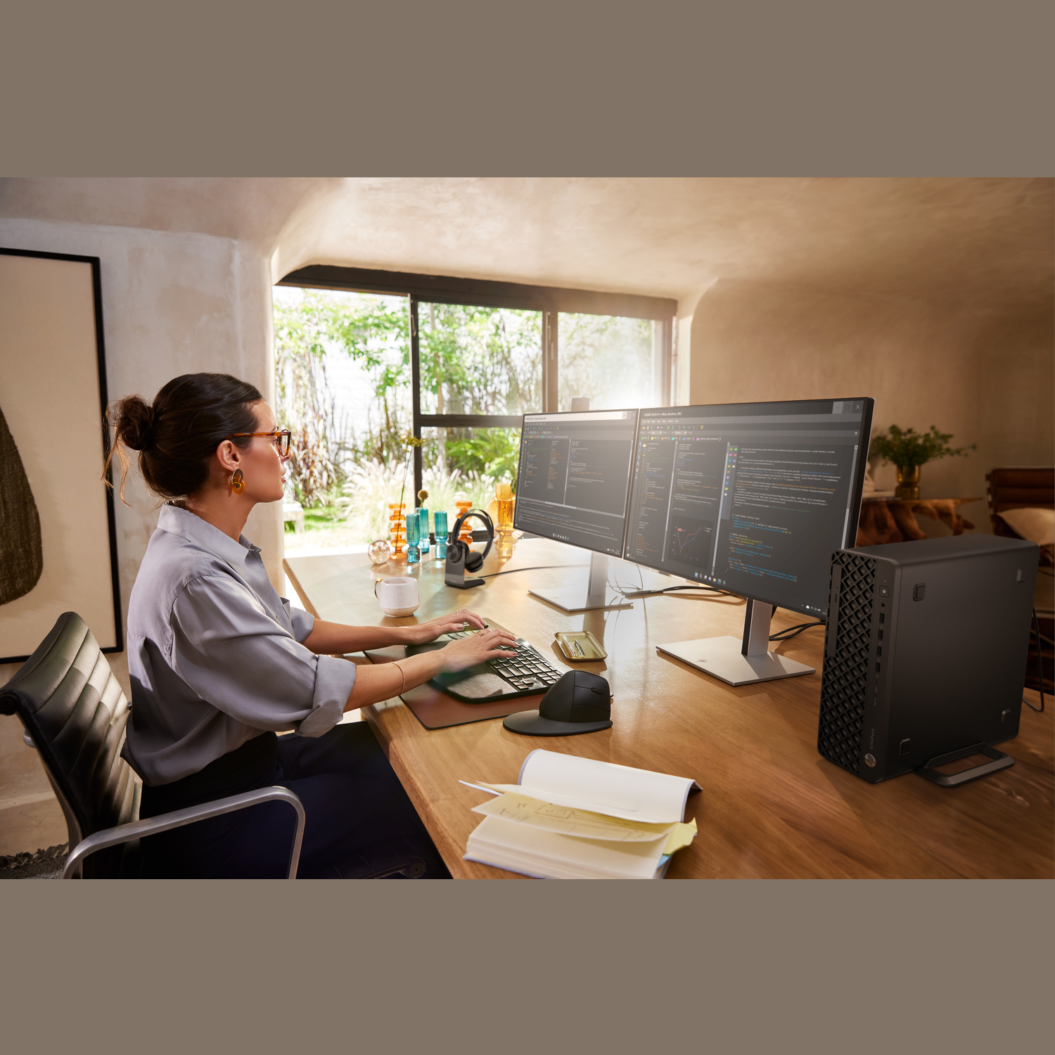 Een vrouw werkt op een computer met twee monitoren. Op het bureau staan een zwarte computer, een toetsenbord en een muis.