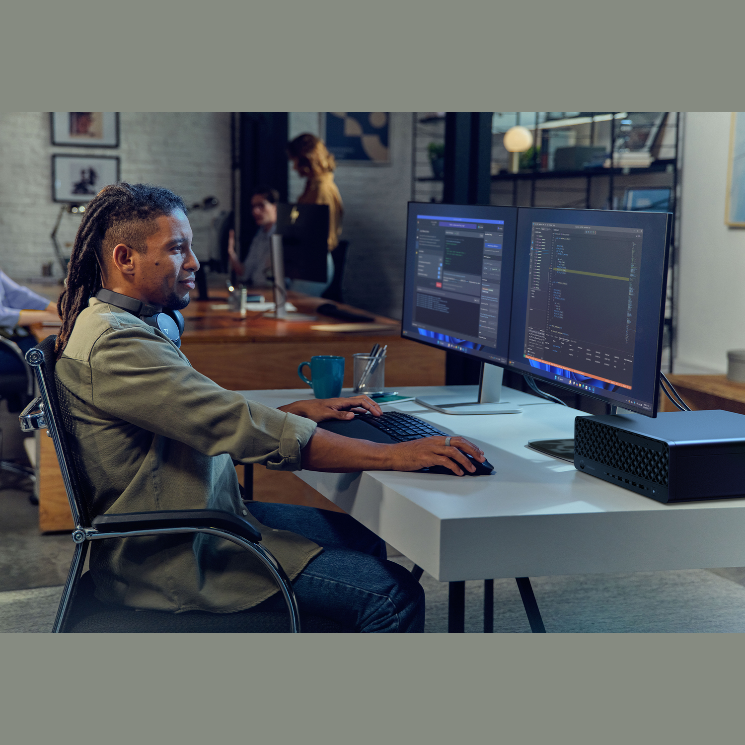 Een man met dreadlocks typt op een toetsenbord voor twee monitoren waarop code wordt weergegeven. Een computer staat op het bureau.
