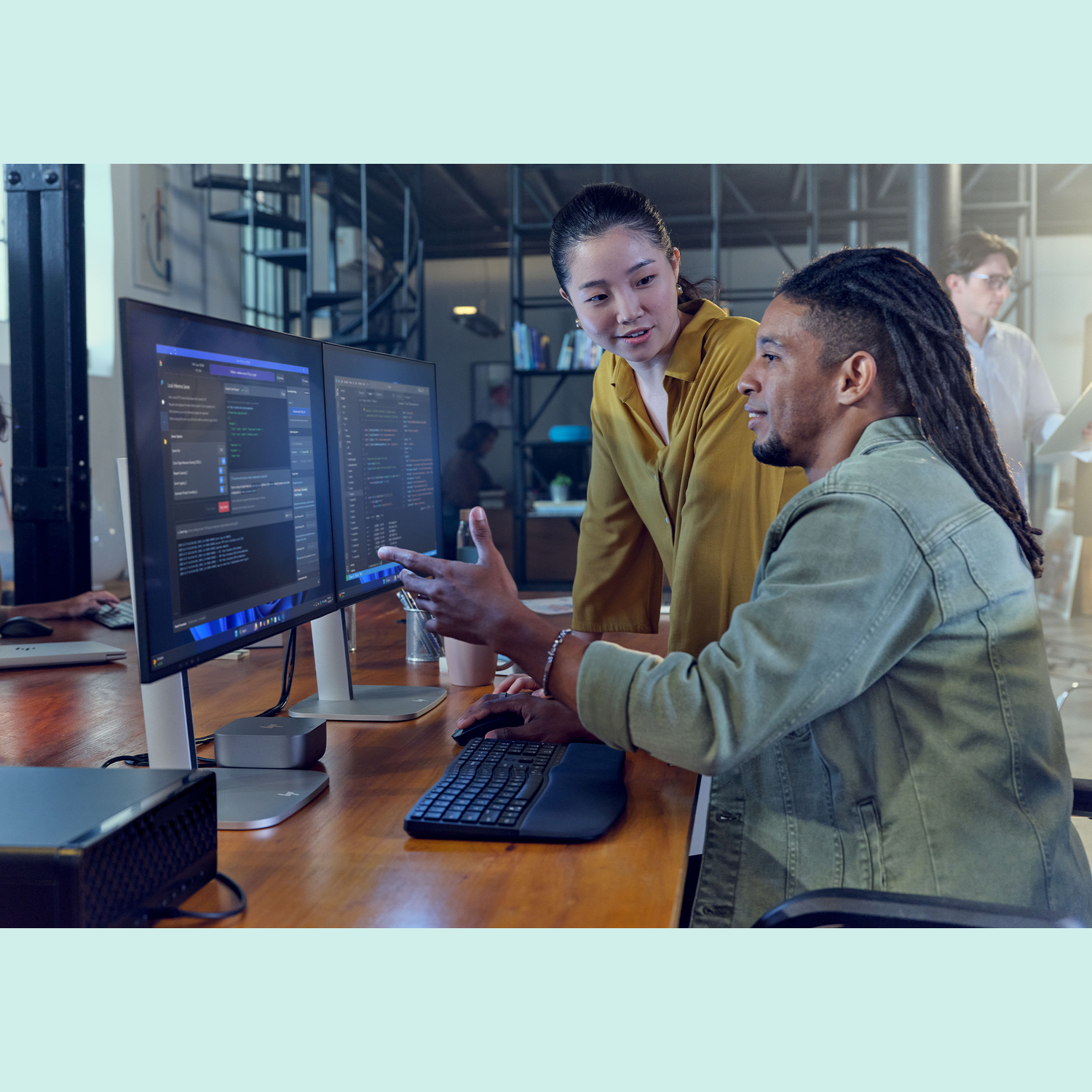Twee mensen kijken naar computerschermen met code. Eén persoon wijst. Anderen werken op de achtergrond.