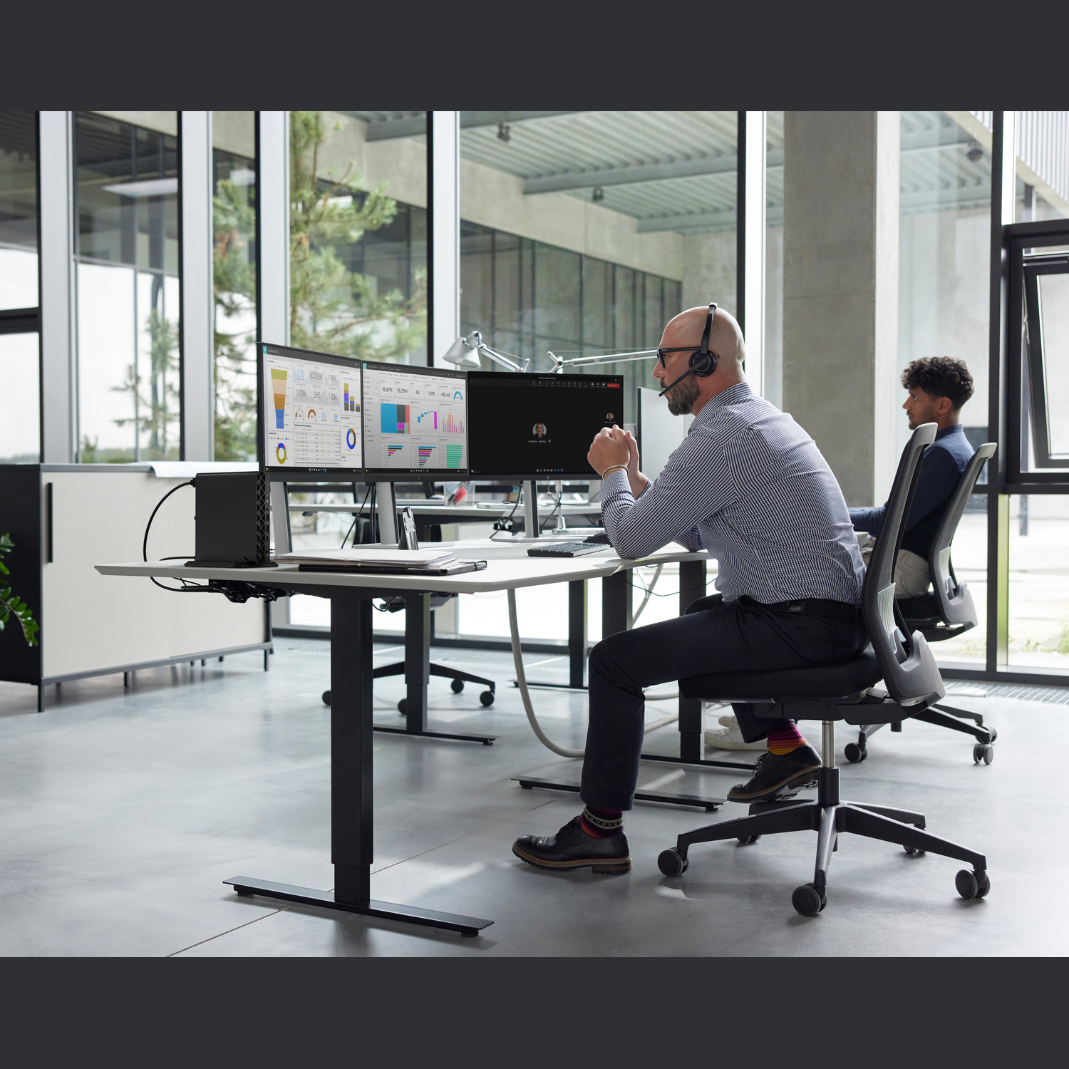 Two men in an office. One wears a headset, looking at a screen with charts. The other sits in the background.