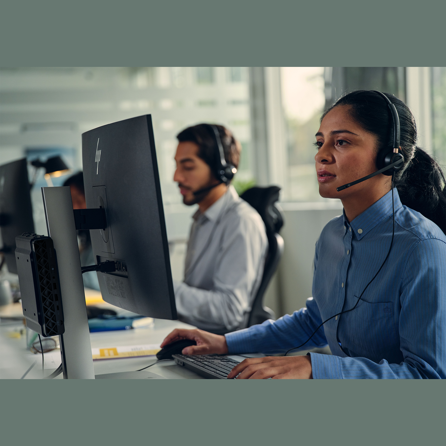 Woman wearing a headset types on a keyboard. A man with a headset is in the background. Both are at computers in an office.