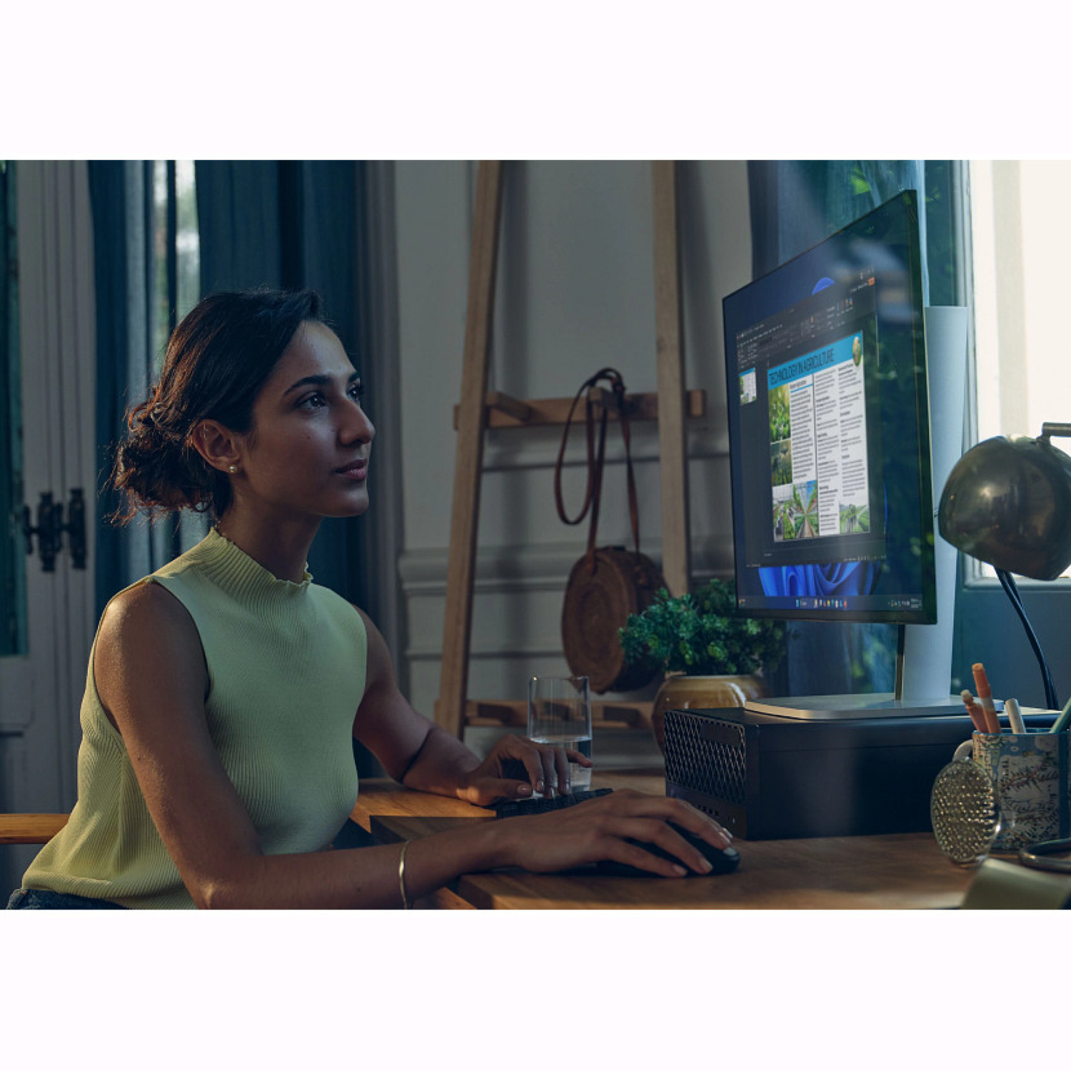 A woman sits at a desk, using a computer. The screen displays an agriculture-related document. A glass of water sits nearby.