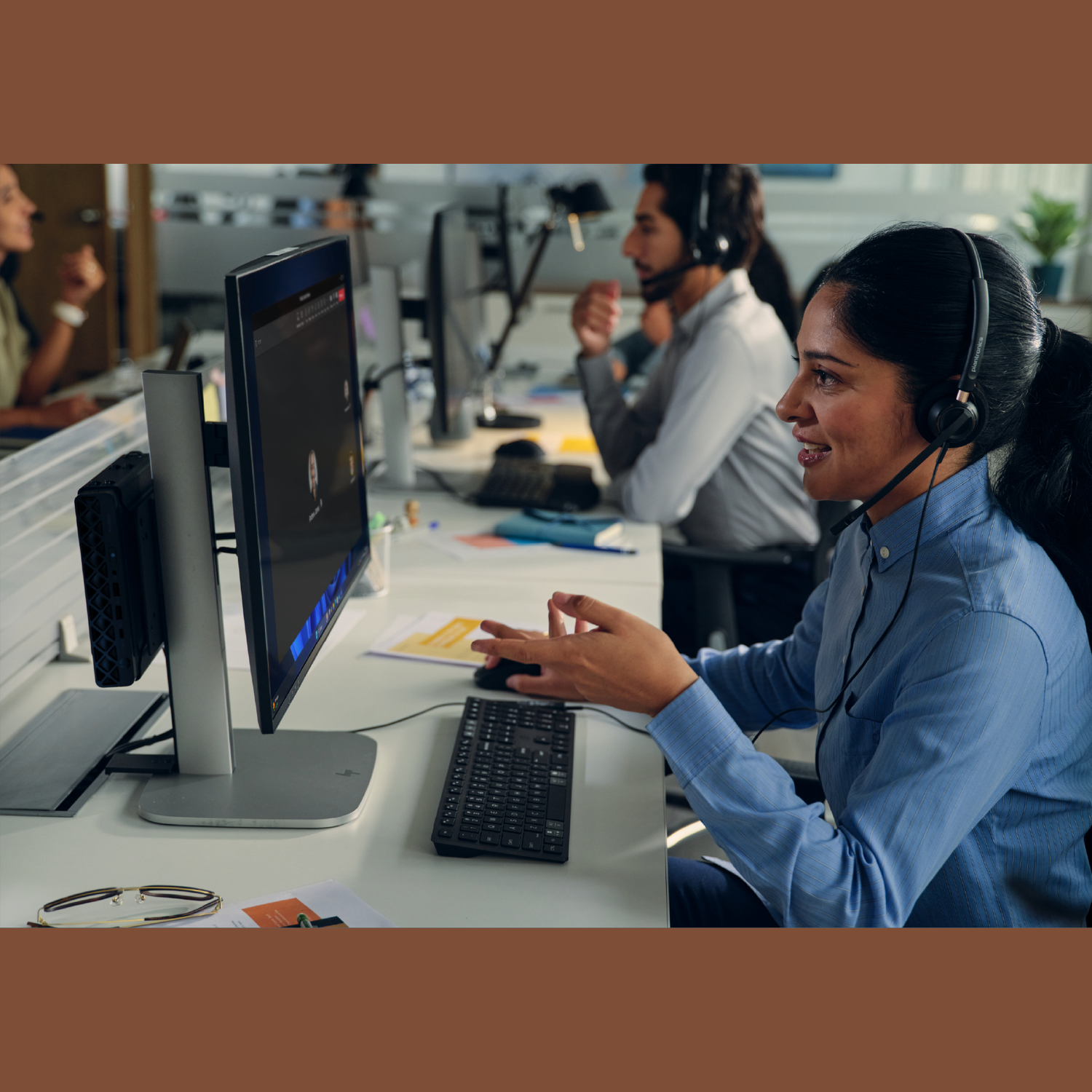 A woman wearing a headset smiles while talking. She is at a desk with a computer. Other people are in the background.