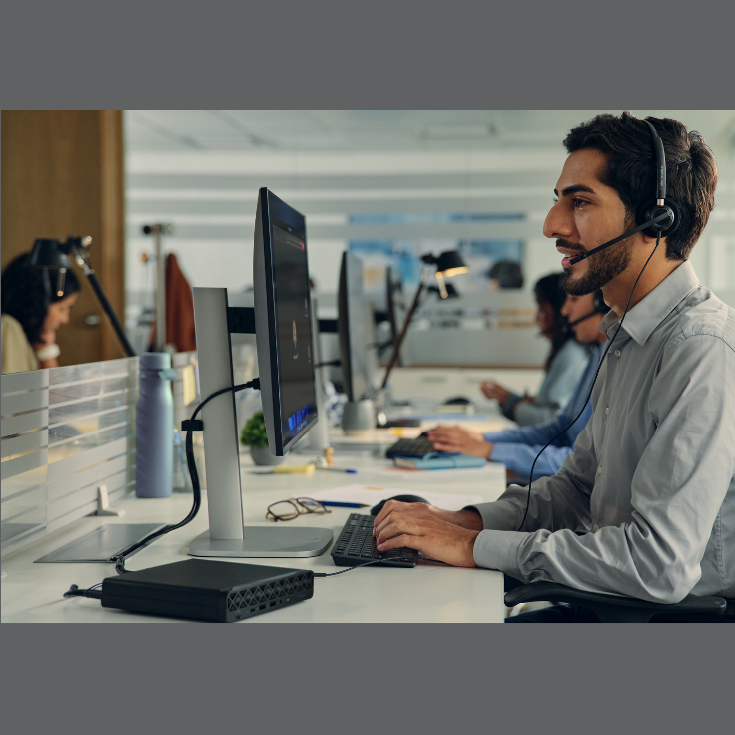 A man wearing a headset and working on a computer in an office. Other people are in the background.