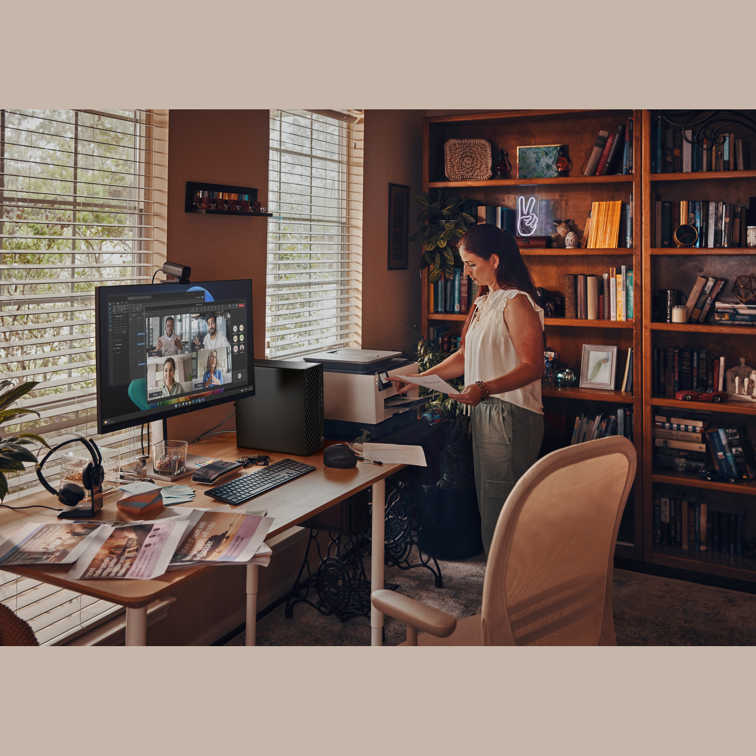 A woman stands at a desk with a computer displaying a video call. Bookshelves and a window are in the background.