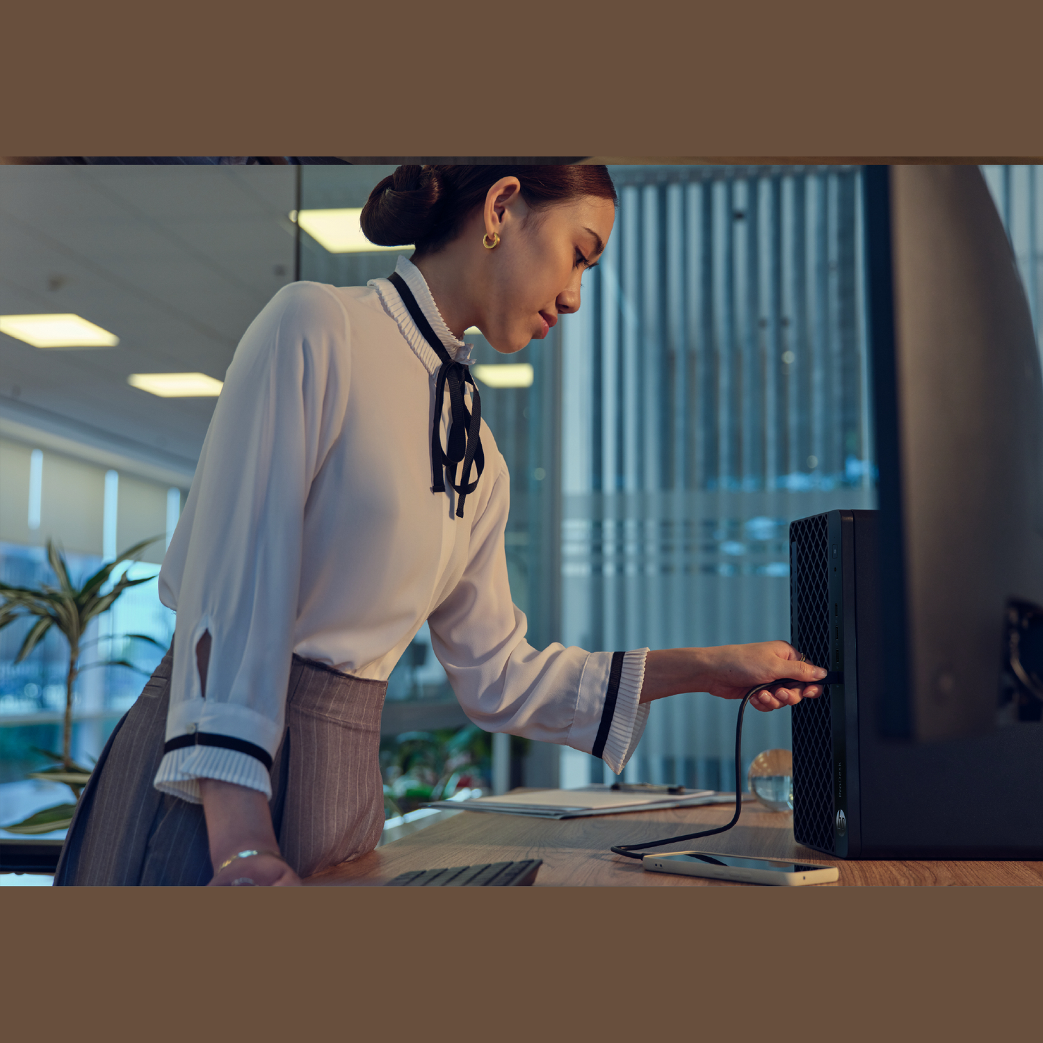 A woman in a white shirt connects a cable to a black computer tower on a desk. A phone and keyboard are also visible.