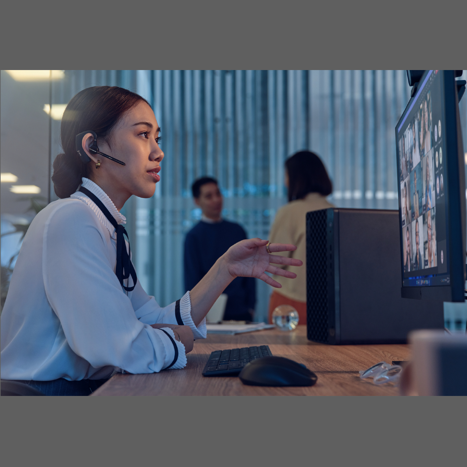 Woman wearing headset gestures during a video call. A desktop computer and two people are in the background.