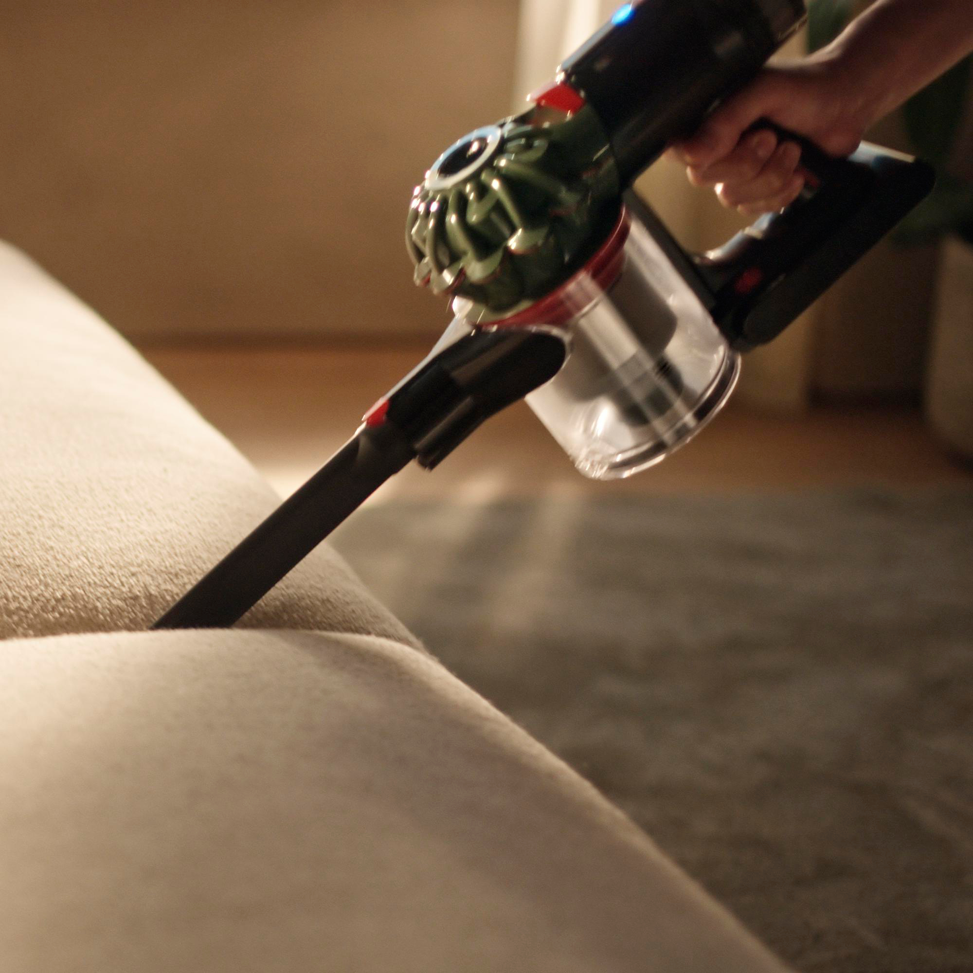 Eine Person, die mit einem Handstaubsauger ein Sofa absaugt. Der Staubsauger ist schwarz und grün.