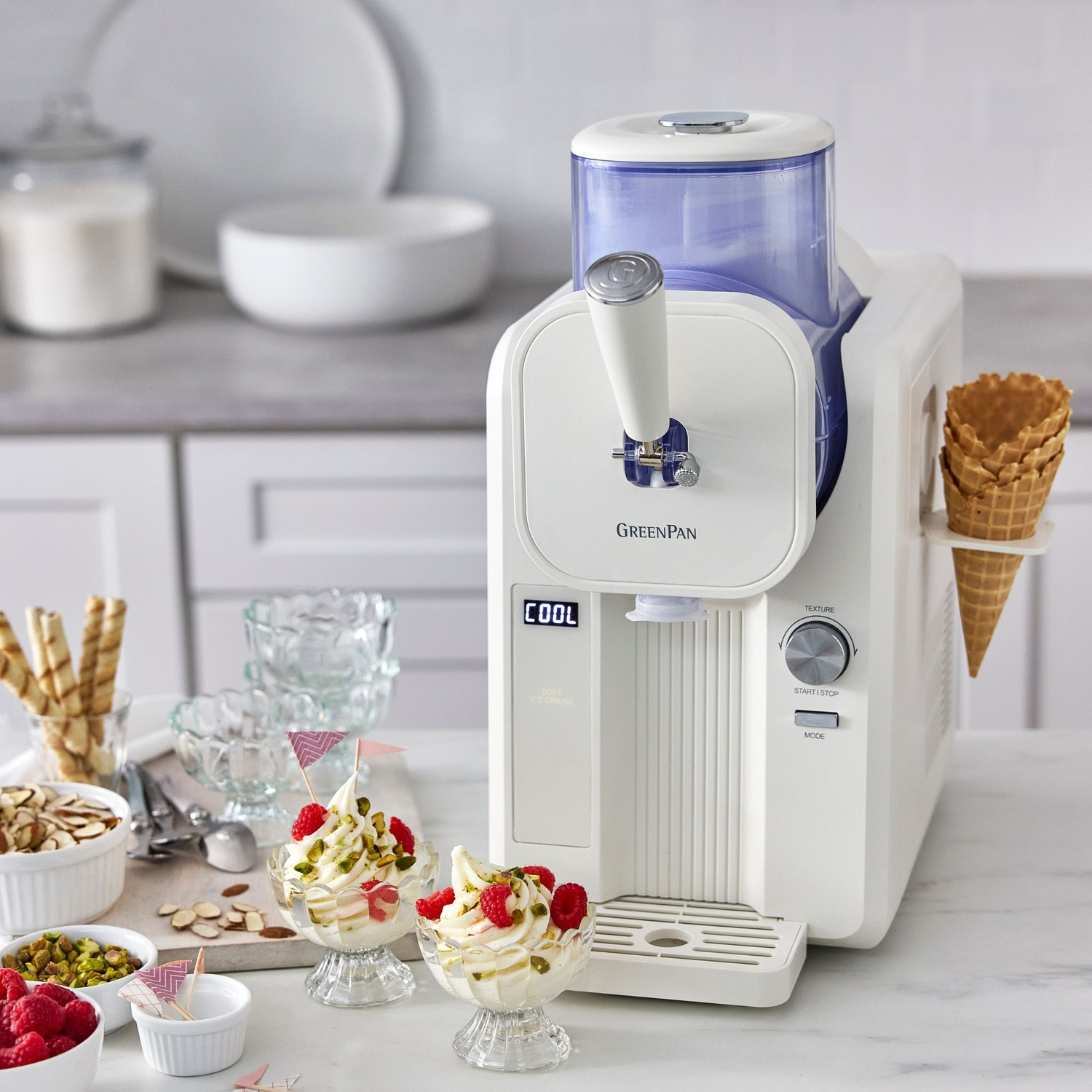 Ice cream maker with two bowls of ice cream, ingredients, and cones. White machine with blue top. Kitchen background.