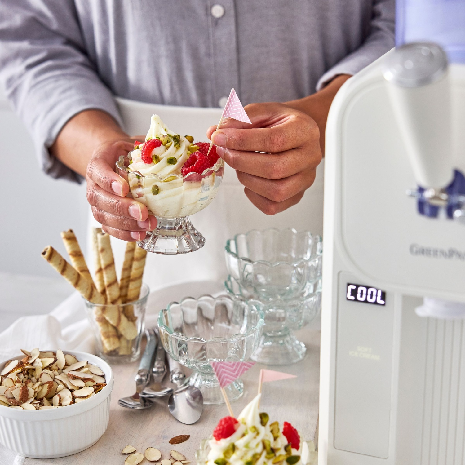 A person decorates ice cream with raspberries and pistachios. Other elements include almonds, wafers, and a soft ice cream machine.