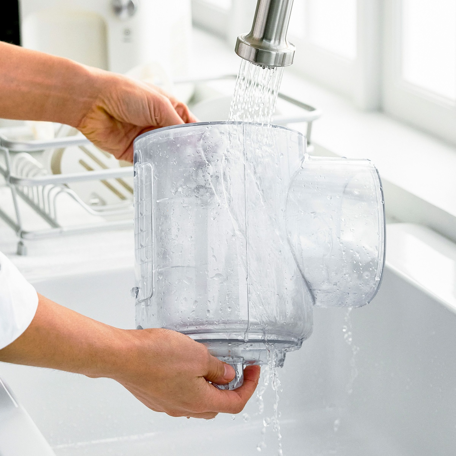Hands washing a clear food processor container under running water in a white sink. Kitchen scene with white background.