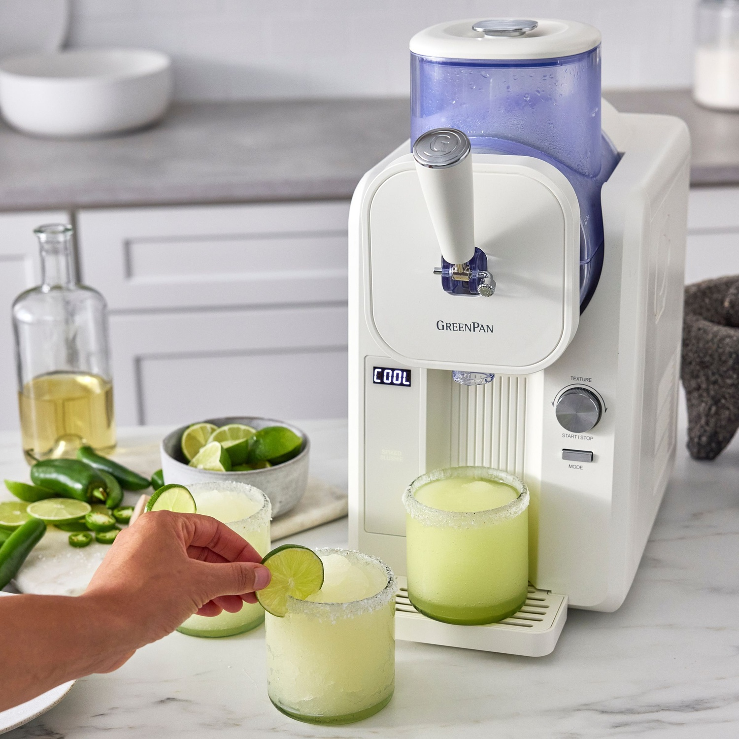 A white margarita machine dispenses drinks. A hand adds a lime slice to a glass. Limes, jalapeños, and tequila are nearby.