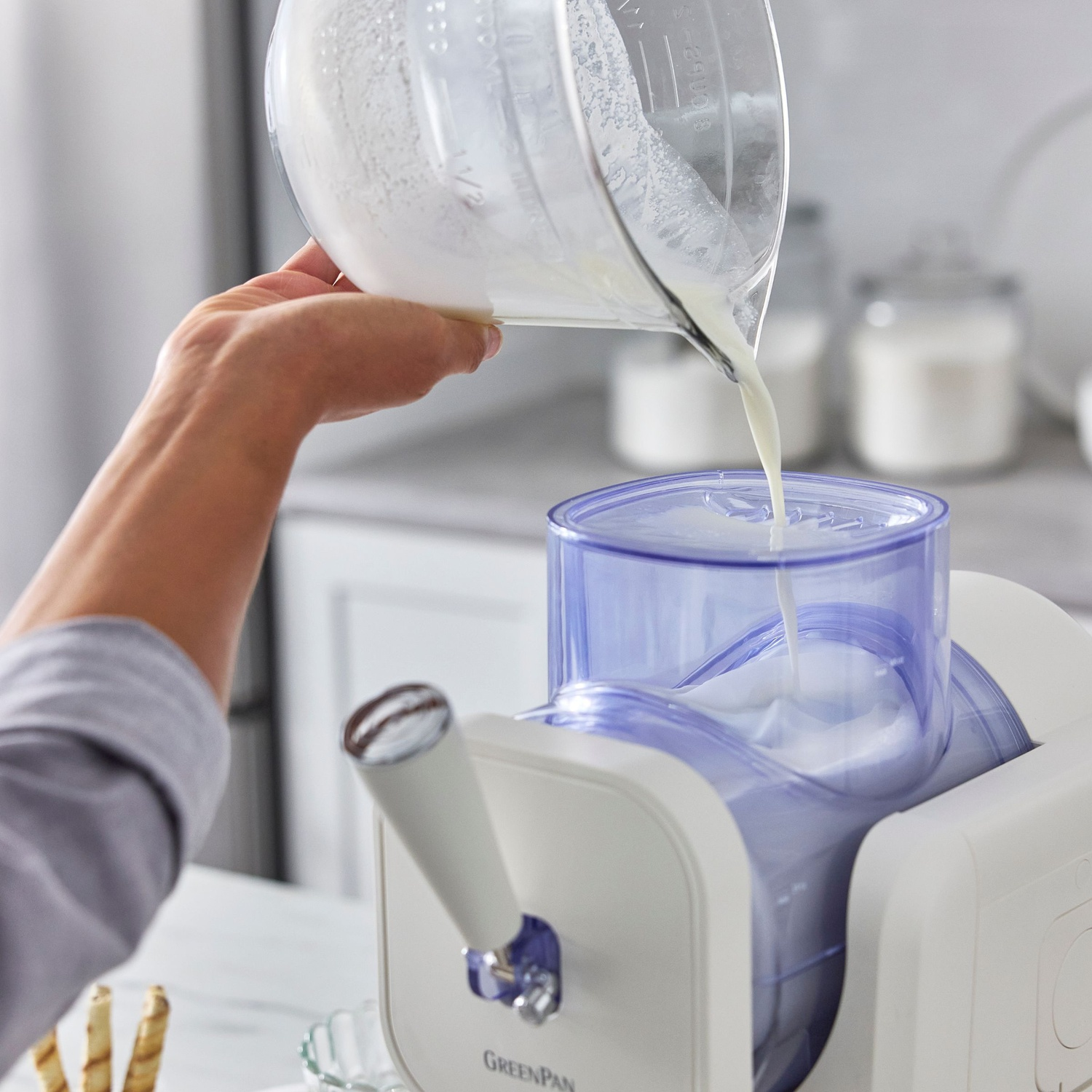 A person pours liquid from a glass measuring cup into an ice cream maker. The maker is white and blue.