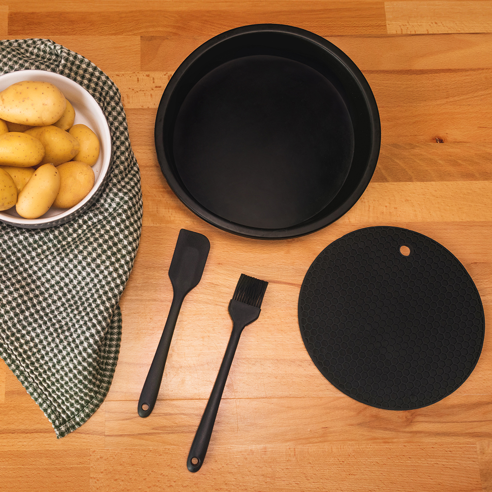 Vista superior de artículos de cocina: patatas en un cuenco, una bandeja negra, espátula, brocha y una alfombrilla redonda sobre madera.