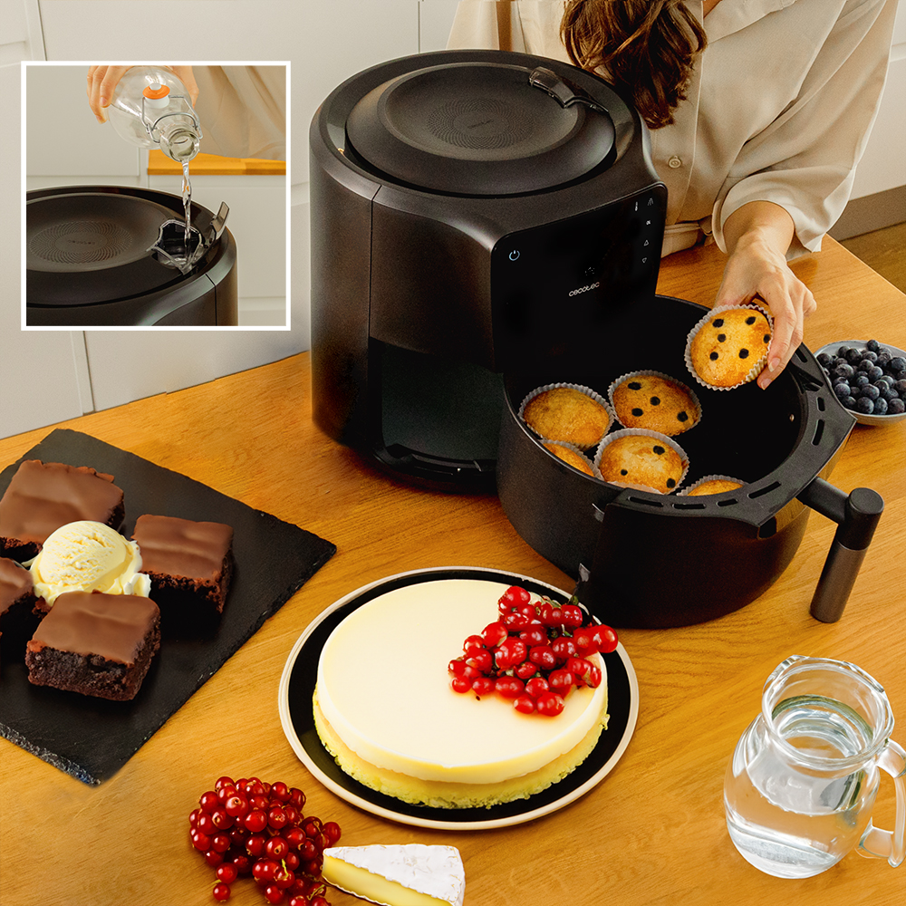 Una freidora de aire con muffins, pastel, brownies y bayas en una mesa de madera. Una persona agrega agua. Una jarra de agua.