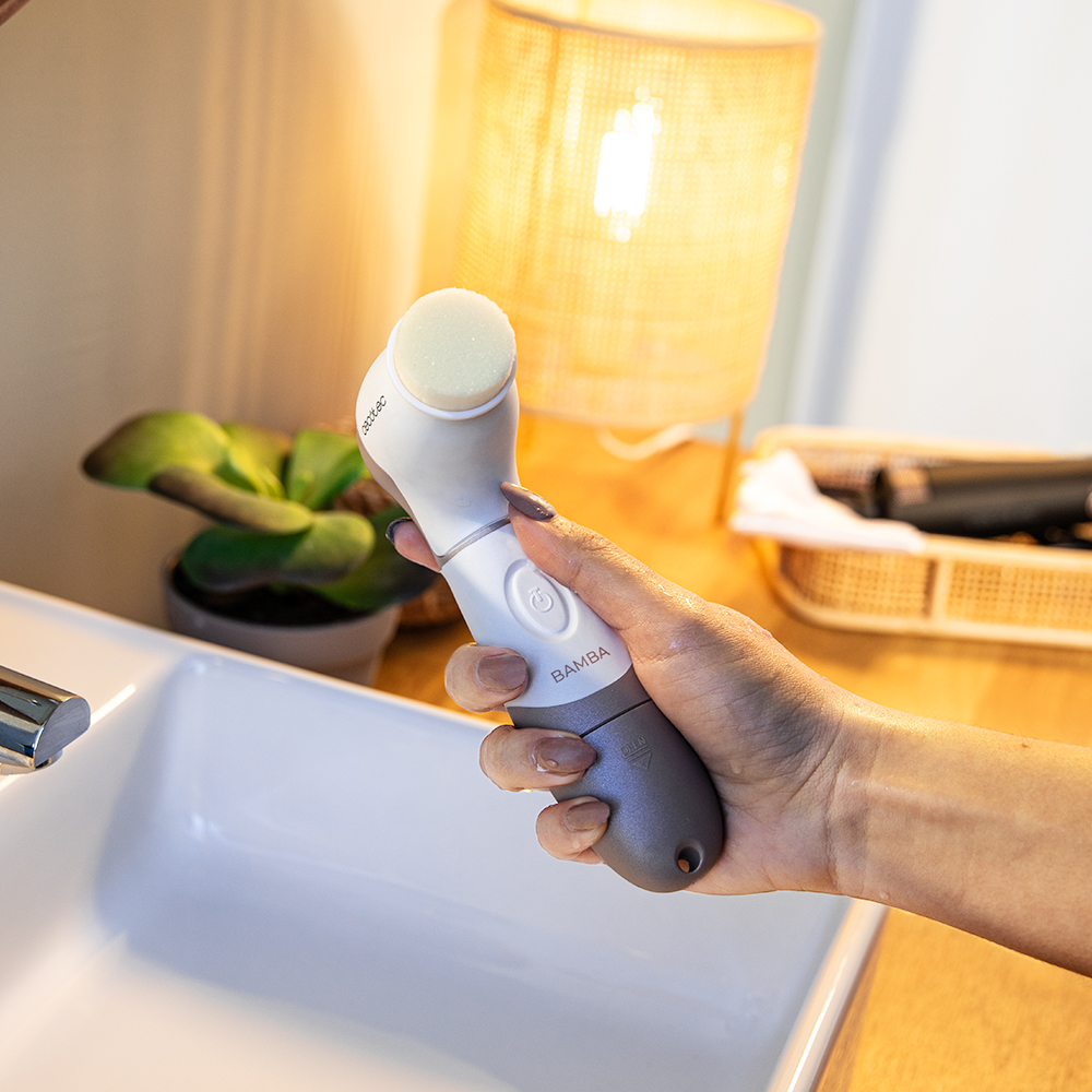 Una mano sostiene un cepillo facial blanco y gris con la etiqueta 'BAMBA' cerca de un lavabo. Una lámpara y una planta están en el fondo.