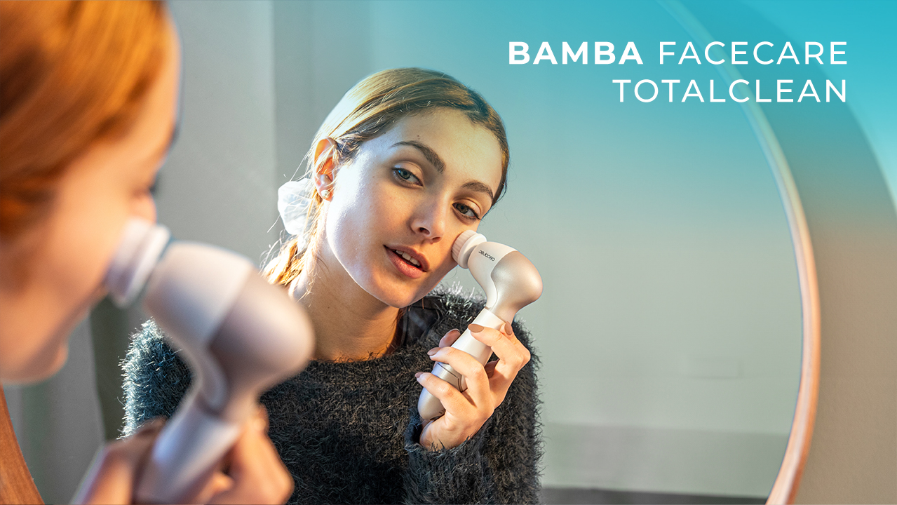 Una mujer usa un cepillo de limpieza facial frente a un espejo. El nombre del producto es visible. Lleva un suéter oscuro.