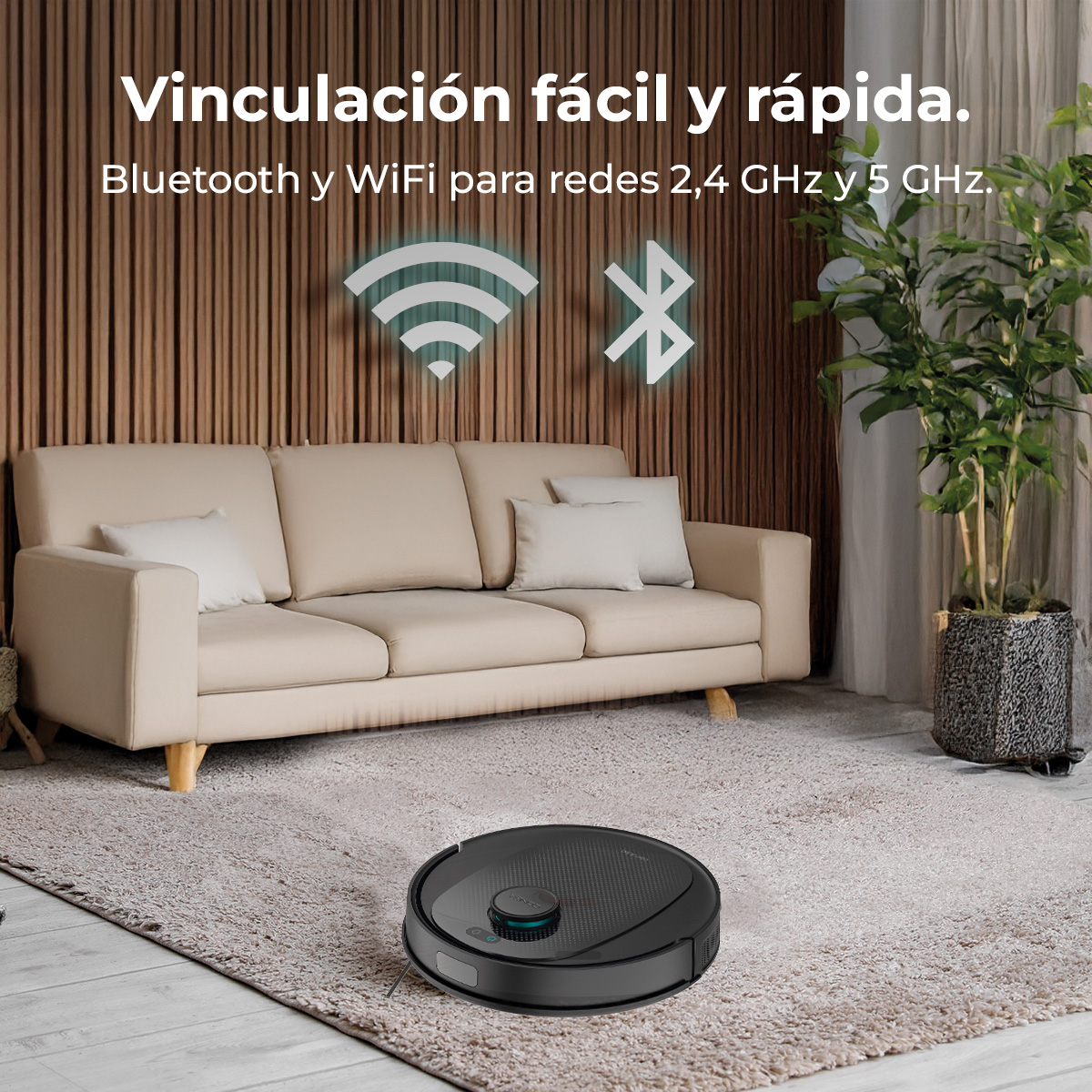 Una aspiradora robot negra sobre una alfombra. Detrás, un sofá beige con cojines blancos. Una planta a la derecha.