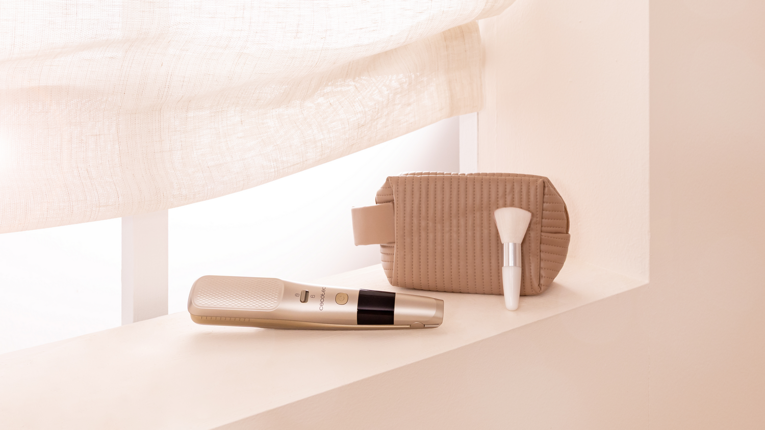 Un set de cosméticos, que incluye un neceser beige, un pincel y un moldeador en un estante. Una cortina beige al fondo.