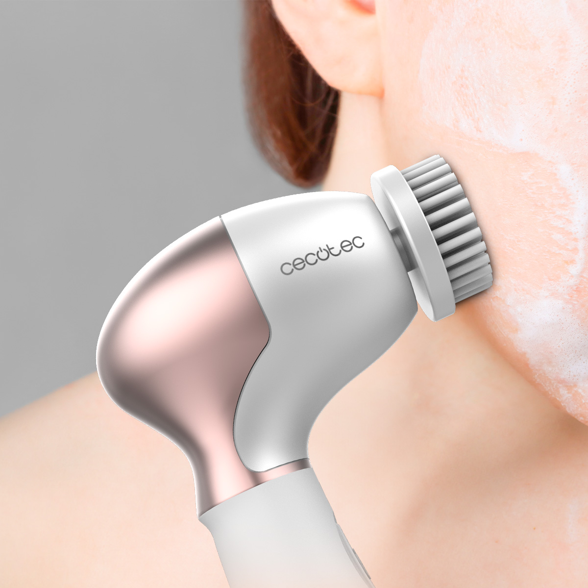 Una persona usa un cepillo de limpieza facial en su rostro. El cepillo es blanco y oro rosa. Jabón espumoso cubre parte de la cara.